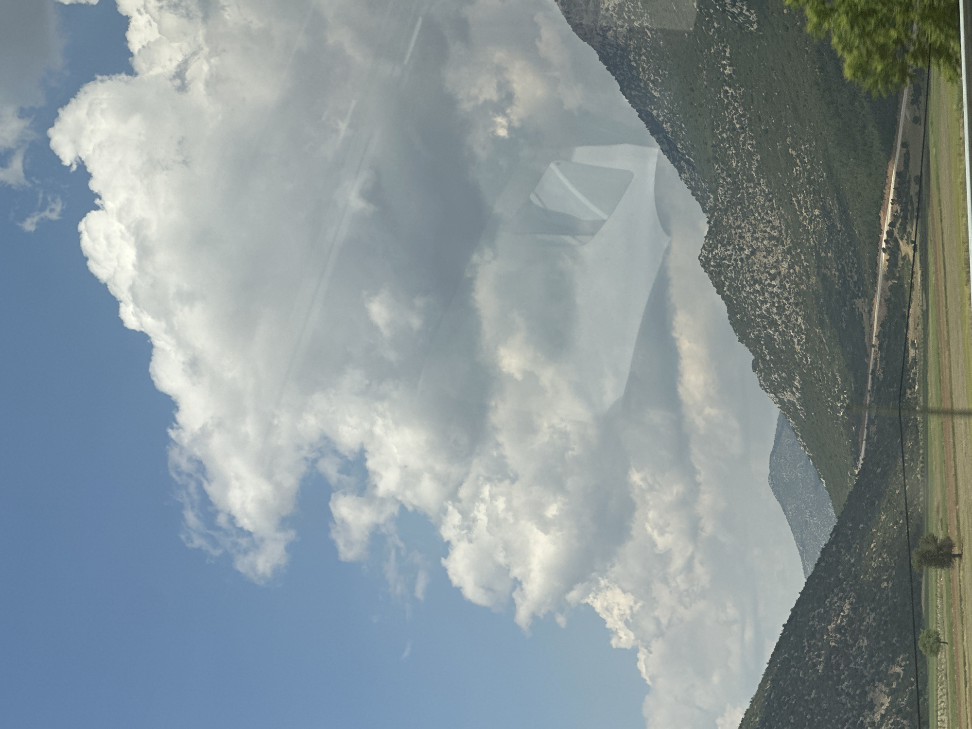The image shows a landscape scene with mountains and clouds. In the foreground, there are green fields with a few scattered trees. A road curves through the foothills of the mountains in the midground. The mountains themselves are covered in green vegetation with rocky outcrops. The sky is blue with large, puffy white clouds. A reflection on the glass, likely from inside a vehicle, shows a partial view of what appears to be a vehicle interior. The scene is consistent with the natural landscape found in and around Korkuteli, Turkey, a town known for its agricultural plains and surrounding mountains. There are no people visible in the image.