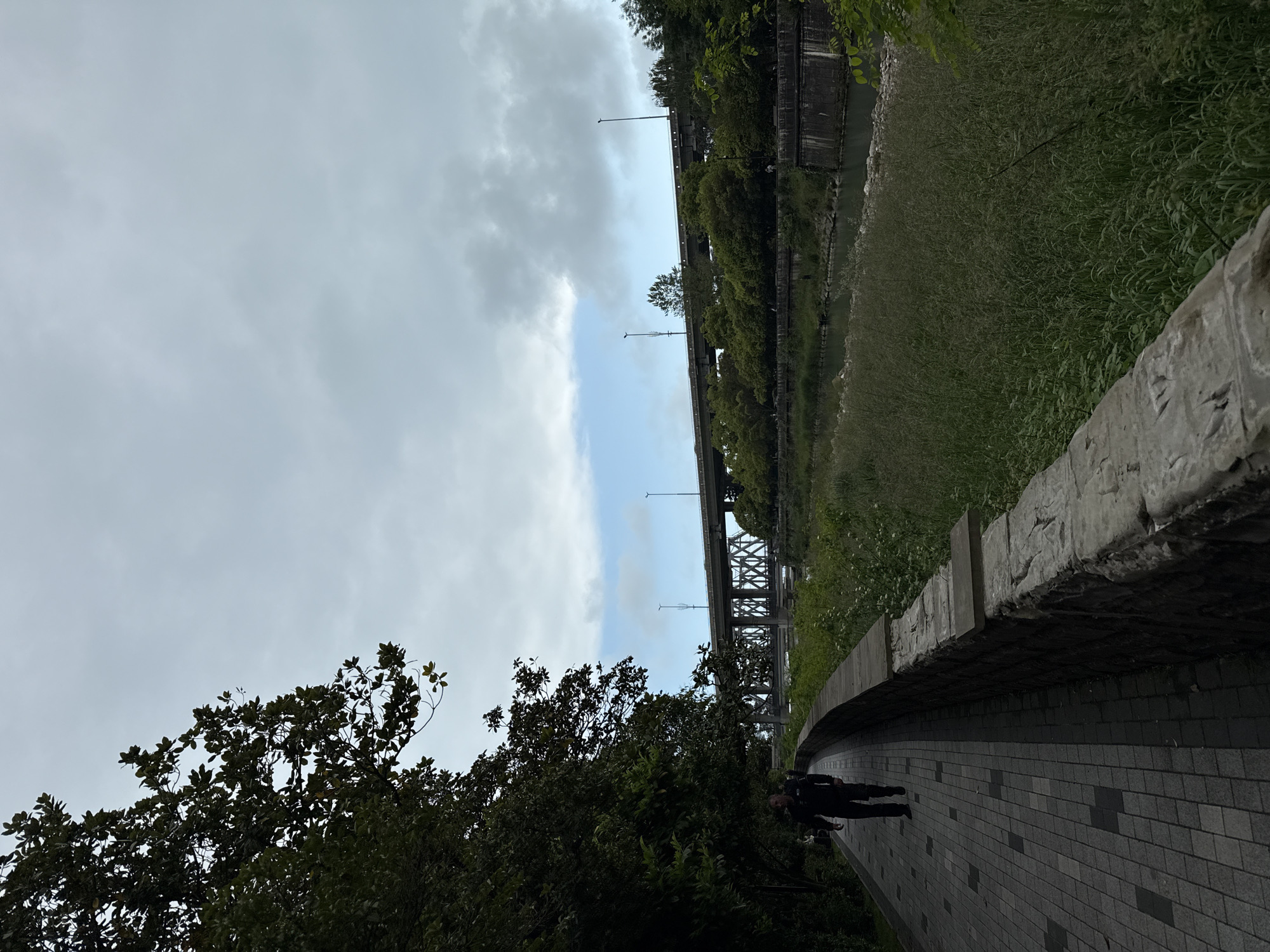 The image captures an outdoor scene in an unknown city. A brick path runs in a slight curve, lined by a low stone wall on the right. To the left of the path is dense greenery. In the background, a bridge spans what appears to be a narrow river or canal, flanked by more greenery on either side. Overhead, the sky is overcast with patches of blue. A person is walking along the path.