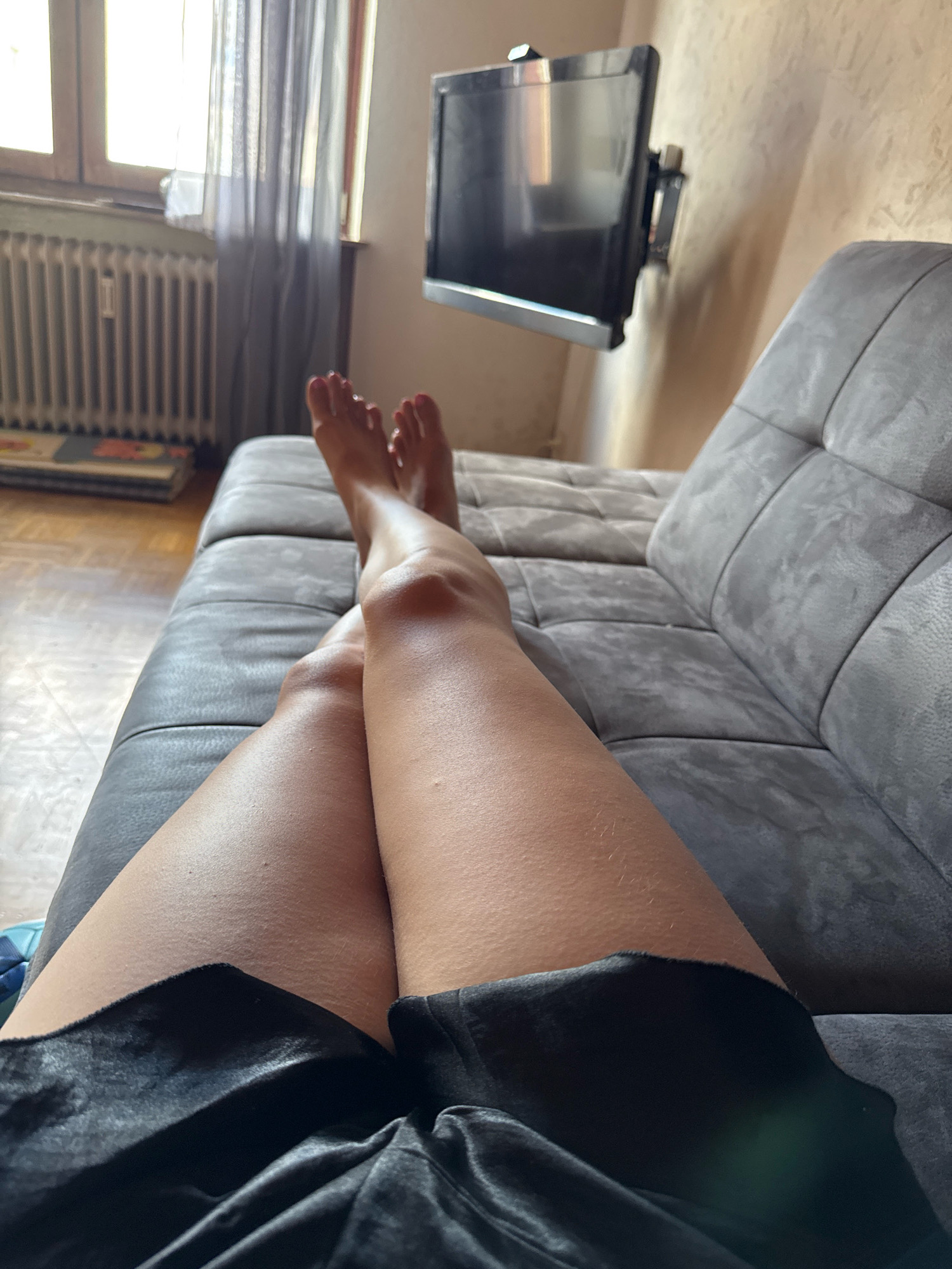 In Überherrn, Germany, a person lies on a grey sofa, their legs and feet visible in the foreground. The person is wearing black shorts. Their feet are bare and positioned towards the upper left of the frame. To the left, a window with grey curtains and a radiator can be seen. A black television is mounted on the wall above and to the right of the sofa. The overall environment suggests an indoor residential setting.