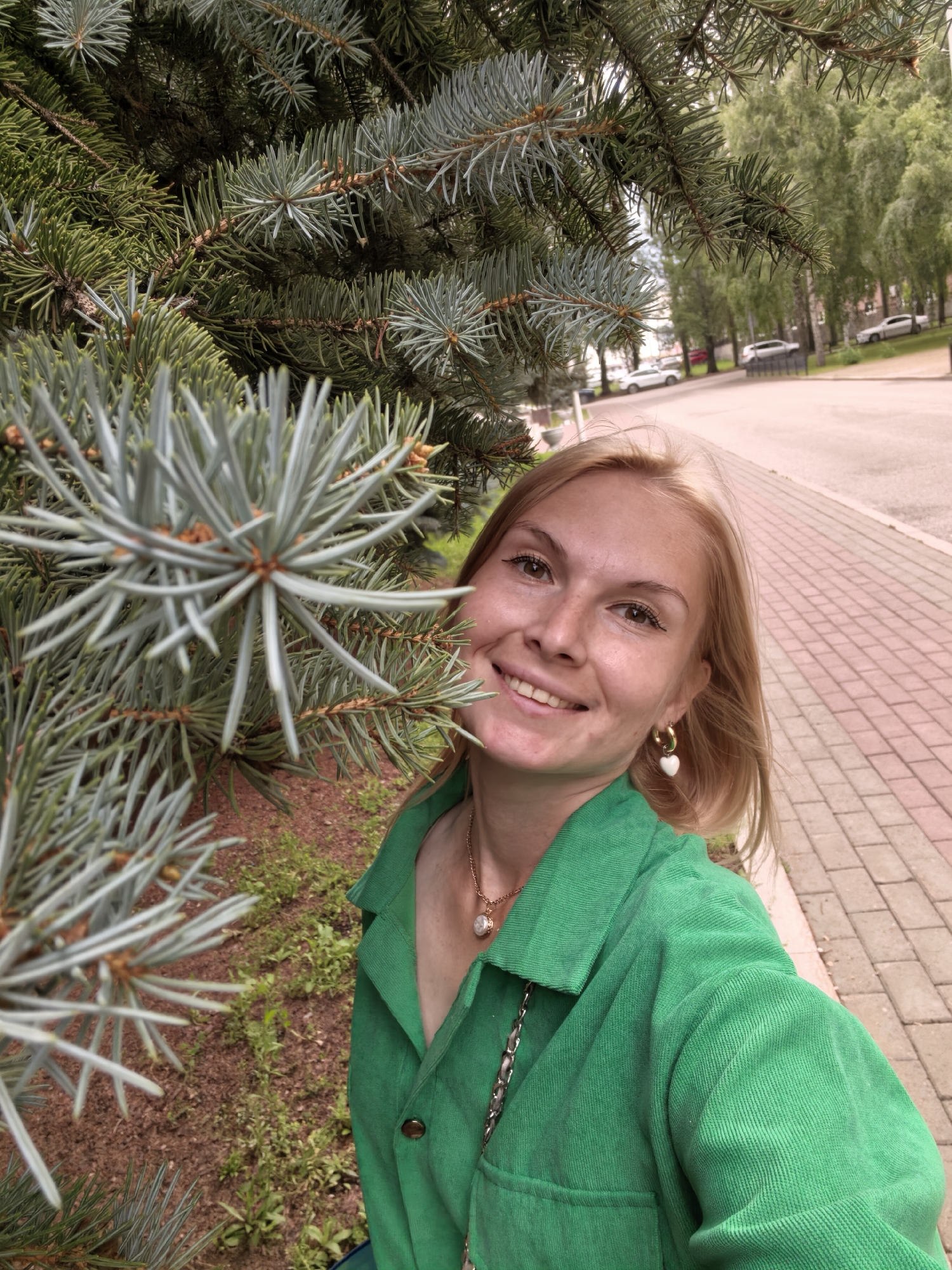 The image is a portrait-style selfie of a young woman with blonde hair and fair skin. She is smiling and looking at the camera. She is wearing a bright green corduroy jacket with a gold necklace that has a pearl pendant. She also has gold hoop earrings, one of which has a white heart charm. The woman is standing outdoors, with a large blue spruce tree in the foreground on the left side of the frame. The tree's needles are a prominent feature, with some in sharp focus and others blurred. Behind her, a paved walkway made of pinkish-red bricks is visible, with a street and parked cars in the background. Trees line the street, suggesting an urban or suburban setting. The lighting suggests it is daytime, likely overcast or late afternoon, as there are no harsh shadows. The overall mood of the image is cheerful and relaxed. The location context provided is Sterlitamak, Russia.
