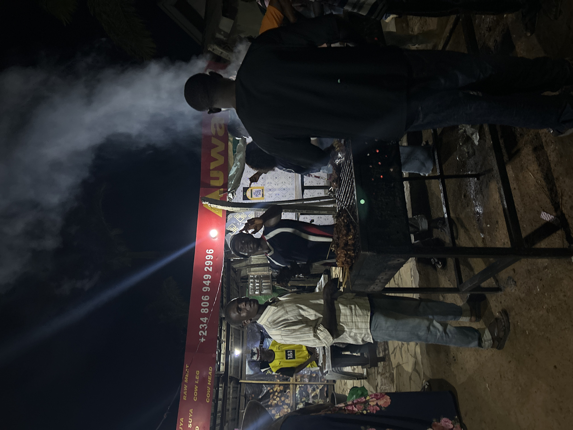 The image depicts an outdoor night scene at a food stall in Bar Jirgi Summa, Nigeria, with a focus on a barbecue operation. Two men are actively engaged in preparing or attending to the food being grilled. One man, wearing a light-colored plaid shirt and blue jeans, stands in front of a large barbecue grill laden with skewers of meat, looking towards the camera. Another man, wearing a dark t-shirt and dark pants, stands behind the grill, facing away from the camera, with smoke rising around him. A third man, wearing a yellow and black sports jersey, is visible in the background, appearing to interact with a customer or colleague. A red banner above the stall displays text, including a phone number, "+234 806 949 2996", and the word "AUWA" is prominently displayed. The ground is dirt, with some scattered debris. The overall atmosphere suggests a busy, local eatery at night.