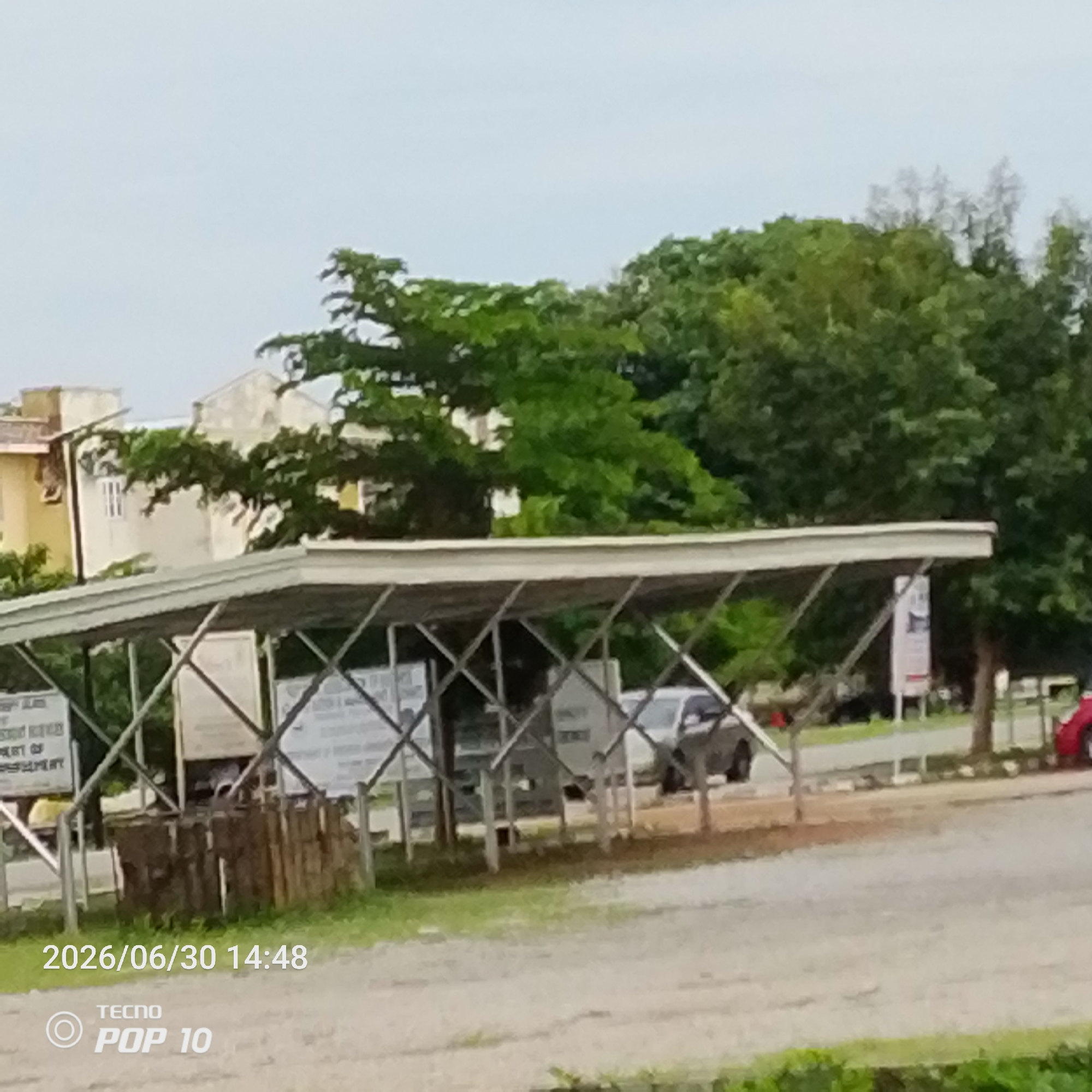 The image depicts an outdoor scene under a cloudy sky, likely in Jalingo, Nigeria. In the foreground, there is a covered parking area with a metal frame and a silver roof. Several signs are visible beneath the shelter, though their text is largely illegible due to image quality and distance. One sign on the left appears to mention "DEPARTMENT OF".
Beyond the shelter, buildings with light-colored walls and a few trees are visible. The setting suggests a public or commercial area. A gray car is parked under the shelter, and a red car is partially visible on the right side of the frame. The ground is a mix of gravel and grass.
The timestamp at the bottom left corner reads "2026/06/30 14:48", indicating the photo was taken on June 30, 2026, at 2:48 PM. The camera model "TECNO POP 10" is also watermarked on the image. The overall atmosphere is ordinary, with no discernible emotions conveyed. The weather appears overcast, contributing to a diffused light.