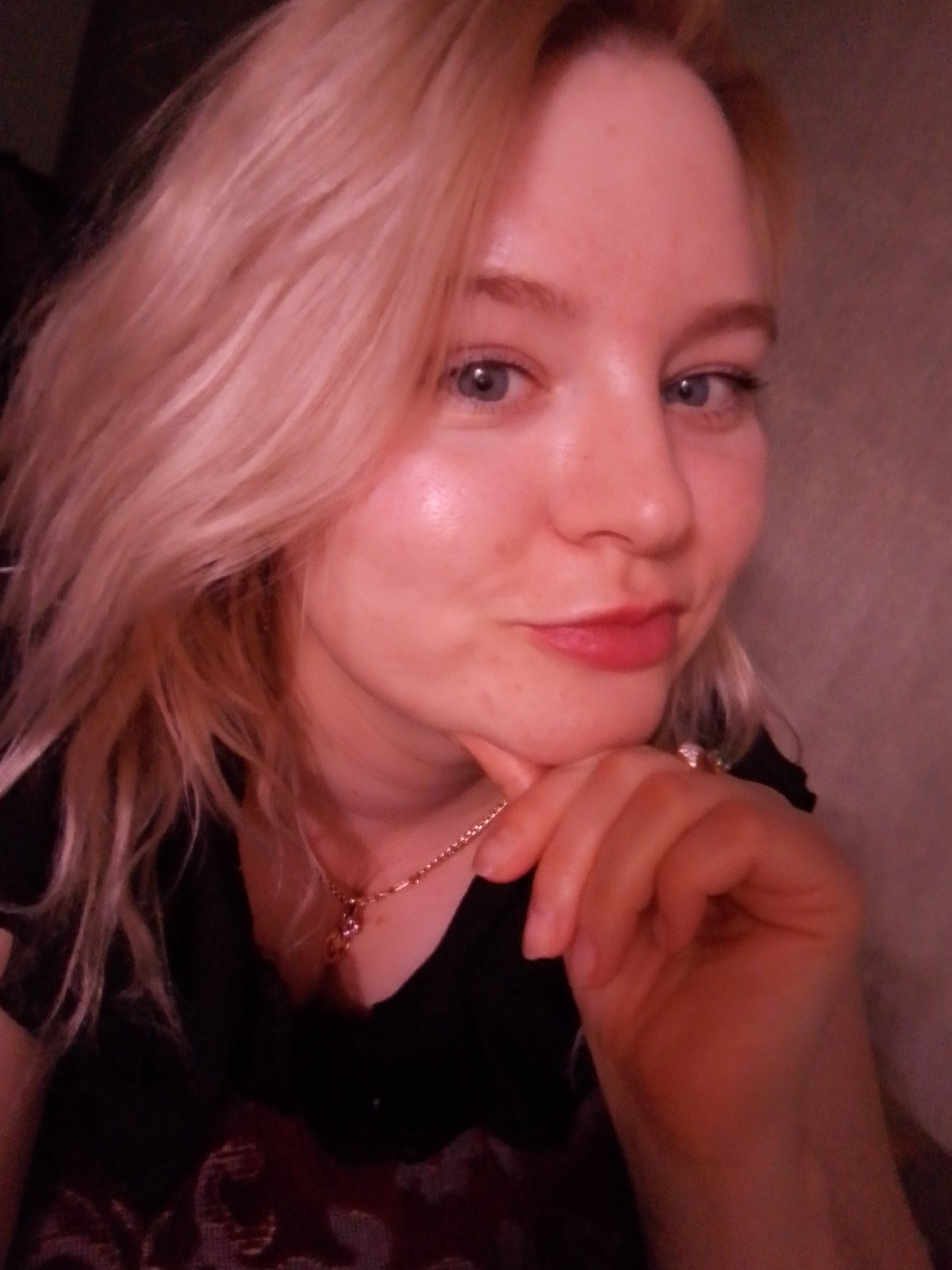 A close-up portrait depicts a single female individual with blonde hair and blue eyes. Her fair complexion shows a slight sheen on the forehead and minor blemishes on the chin. She has full, reddish-tinted lips and a slight smirk. She wears a black top, partially revealing a dark, patterned fabric below. A gold-colored chain with a small pink or purple stone pendant rests on her chest. A ring with a clear or white stone is visible on the ring finger of her right hand. Her right hand is positioned with curled fingers resting against her chin. The individual is looking directly at the camera. The background consists of a textured, light brown or beige wall to the right, with an indistinct dark area to the left. The scene is illuminated by soft, warm-toned light, indicating an indoor setting in Izhevsk, Russia.