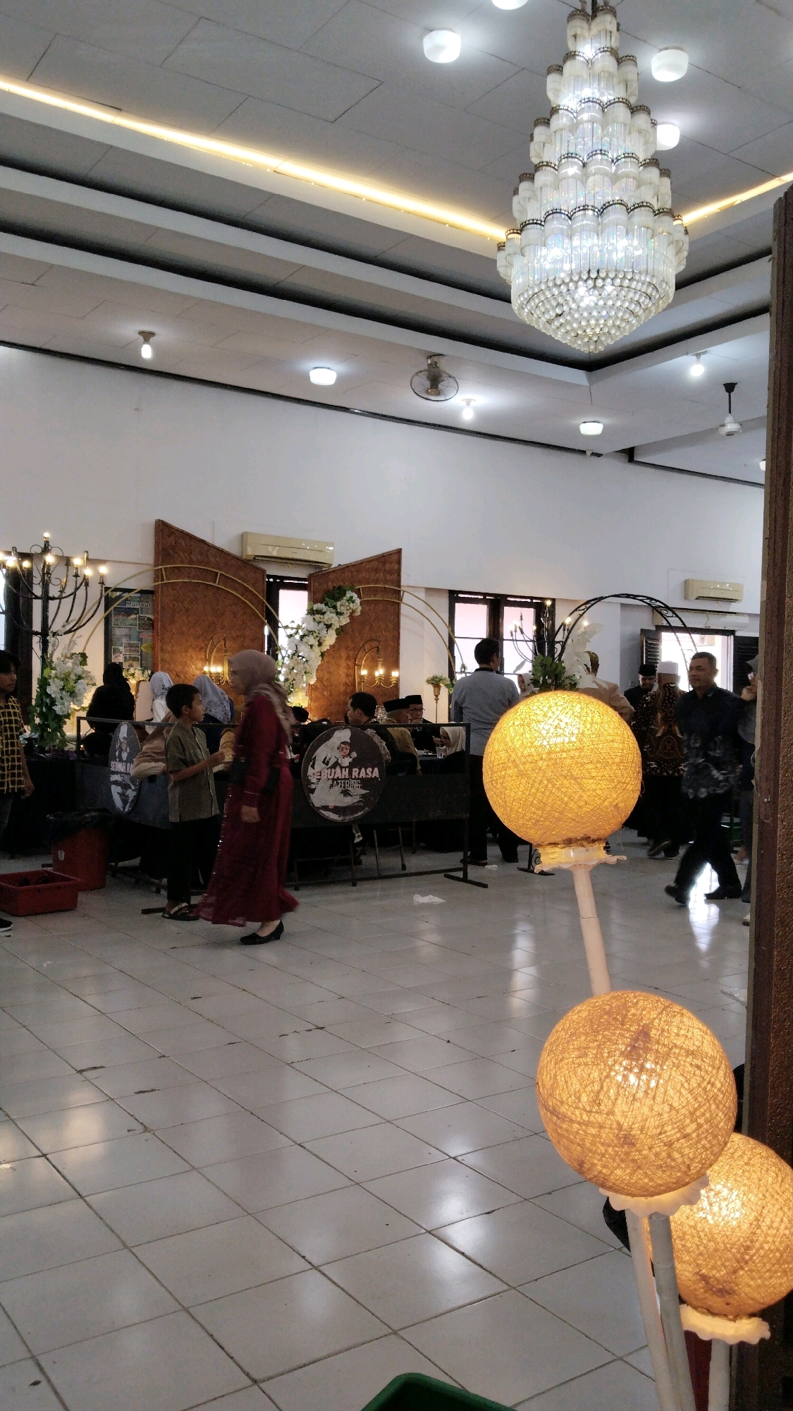 The image depicts an indoor event likely taking place in Jember, Indonesia, possibly a wedding reception or a formal gathering, given the chandelier and decorative elements. The environment appears to be a function hall or a similarly sized space with white tiled floors and white walls. The ceiling features a prominent, ornate chandelier with multiple tiers of crystal-like elements, and there is a strip of warm-toned lighting running along a section of the ceiling.
In the foreground, two spherical lamps with a woven texture, illuminated from within with a warm yellow light, stand on poles. The lamp in the front is closer to the camera, while the second is slightly behind and to the right.
Several people are visible in the midground and background. On the left, a young boy in a light green shirt is facing a woman in a dark red dress, who is walking away from the camera. Further back, a group of people are seated at tables, some dressed in traditional Indonesian attire like batik shirts and headscarves. A decorative backdrop featuring wooden panels and white floral arrangements is set up behind these tables.
On the right side of the image, a man in a dark patterned shirt and pants is walking away from the camera, and other individuals are present in the background, suggesting ongoing activity within the venue. Some of the tables appear to have circular signs with images and text, possibly related to catering services or event sponsors. The overall atmosphere suggests a lively event with people mingling and participating in the occasion.