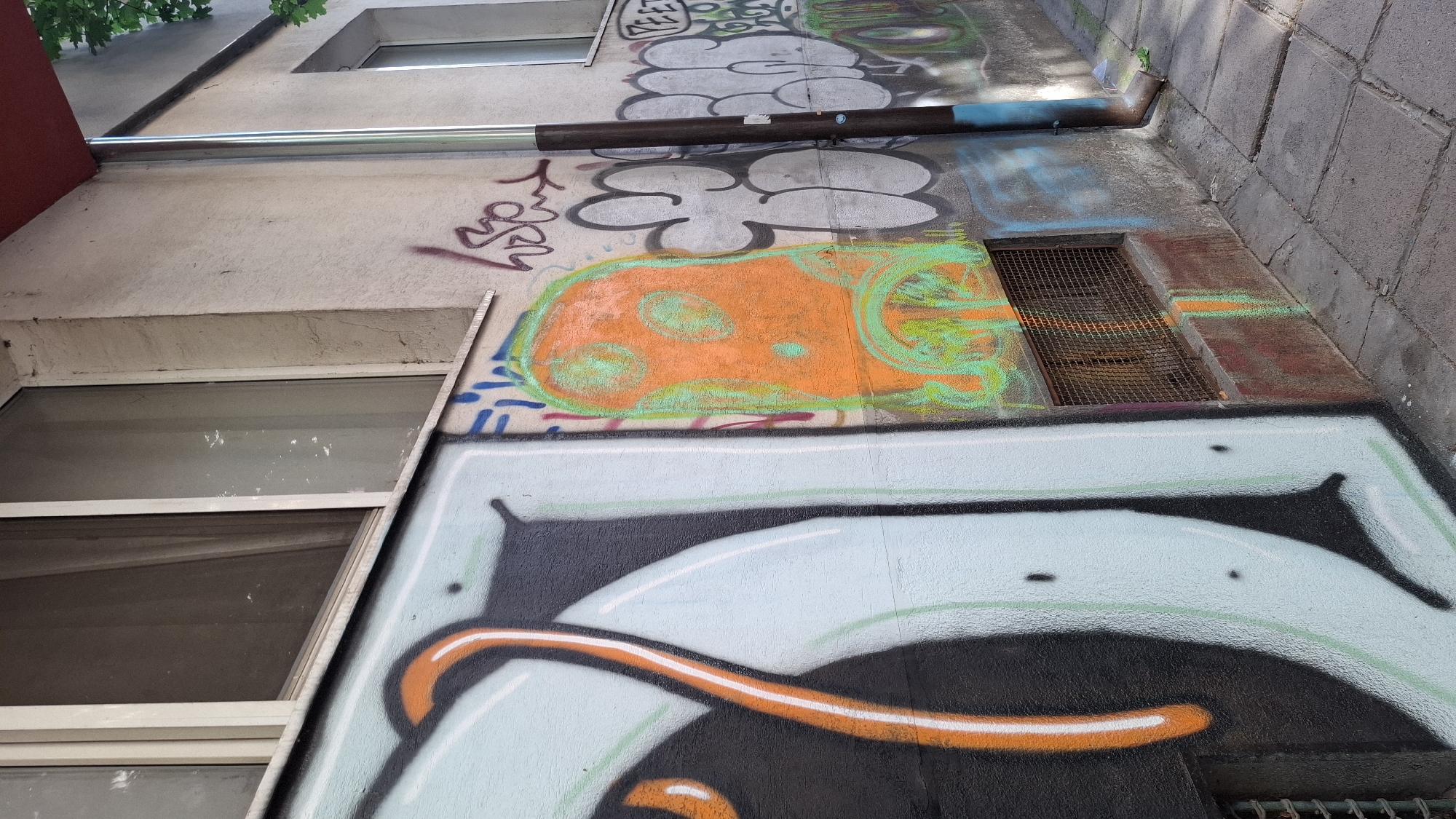 The image depicts a section of an exterior wall of a building, covered in graffiti, with a window and a downspout visible. The wall is concrete and appears weathered, with cracks in its surface. The graffiti is in various colors and styles, including large, bold letters in light blue and black, an orange and green abstract figure resembling a ghost, and smaller tags and scribbles. A metal grate covers a small opening near the base of the wall. A downspout, made of metal, runs vertically along the wall. The ground in front of the wall is paved with concrete tiles. The scene suggests an urban environment, likely in a city where graffiti art is prevalent. The question implies a specific location within Sofia, Bulgaria, but this information cannot be deduced solely from the visual content of the image. There are no people or animals present in the frame. The overall lighting suggests it is daytime, with sunlight casting shadows on the ground.