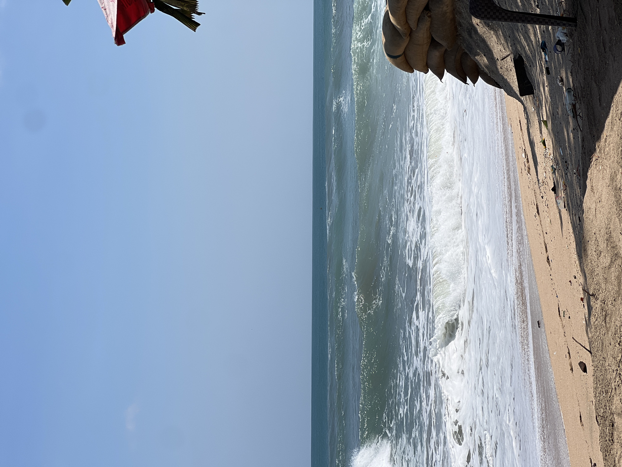 An eye-level, medium shot captures a view of the ocean and a sandy beach on a clear day. In the foreground, the sand is littered with debris, including plastic bottles and wrappers, suggesting a lack of recent cleanup. Several sandbags are piled up on the right side of the frame, possibly for flood protection, and a dark woven chair is partially visible. Beyond the beach, the ocean stretches out, with white-capped waves rolling towards the shore. The water is a deep blue-green, and the horizon is a clear line where the sea meets the pale blue sky. In the upper right corner, a portion of a red umbrella and some palm fronds are visible, indicating a temporary structure or vegetation above the scene. The overall impression is one of a natural coastal environment marred by human-generated waste and possibly protective measures against the elements.