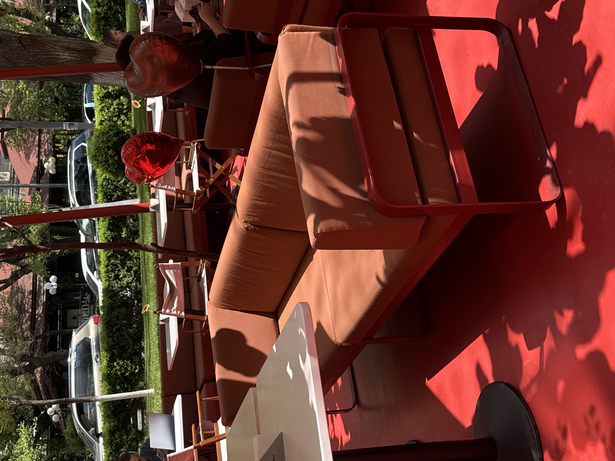 This image captures an outdoor seating area, likely part of a cafe or restaurant patio, bathed in bright sunlight in Yerevan, Armenia. The scene is characterized by a vibrant red ground that contrasts with the lush green trees and bushes in the background.
The primary subjects are the outdoor furniture and a few patrons. The seating comprises comfortable-looking sofas upholstered in an earthy orange-brown fabric, set within matching red metal frames. White outdoor tables are also visible, some with wooden director's chairs alongside them.
A key detail is a woman in the mid-ground, holding two prominent heart-shaped foil balloons: one a deep bronze-red and the other a shiny, bright red. These balloons suggest a celebratory or romantic occasion, adding a festive touch to the relaxed atmosphere. Other people are partially visible, seated on the sofas, enjoying the outdoor setting.
The strong sunlight casts long, dynamic shadows of the furniture and surrounding trees across the red floor, indicating it is likely midday or late afternoon on a clear, sunny day. Beyond the patio, a street scene unfolds with parked cars, including SUVs and sedans, alongside well-maintained hedges and mature trees. A building with a reddish-brown roof and white street lamps are visible in the distance, confirming an urban or suburban outdoor location. The overall impression is one of leisure and enjoyment in a warm, inviting environment. No discernible text is visible.