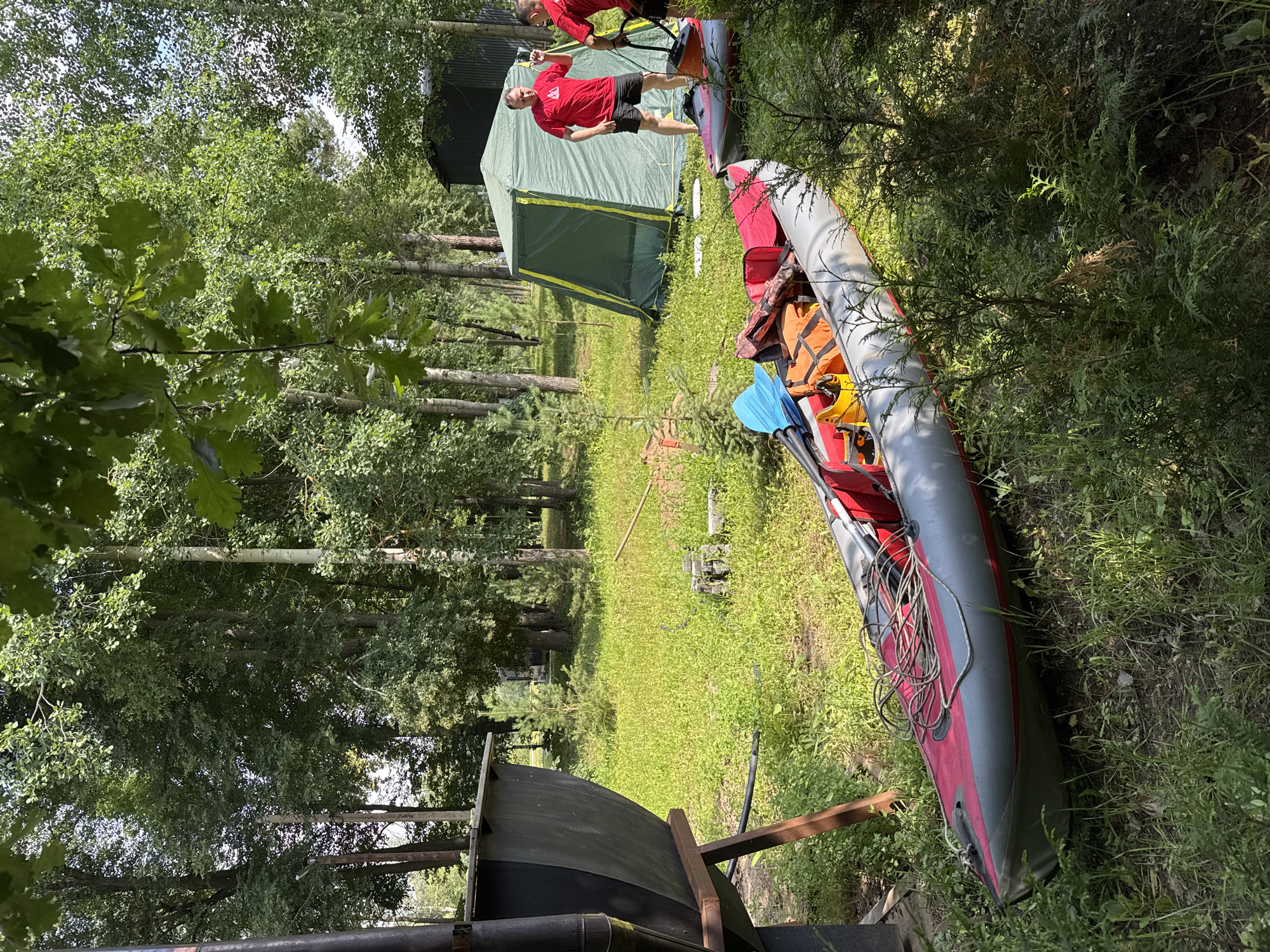 In a wooded area with a grassy clearing, a campsite is set up. Two men, one in a red t-shirt and black shorts, the other partially obscured, stand near a green tent. In the foreground, an inflatable kayak is on the ground, packed with gear including orange bags and a blue paddle. To the left, a large, dark, industrial-looking container sits on a metal stand. The environment suggests a recreational camping and water activity setting. The location is Doma 1096 Km, Russia.