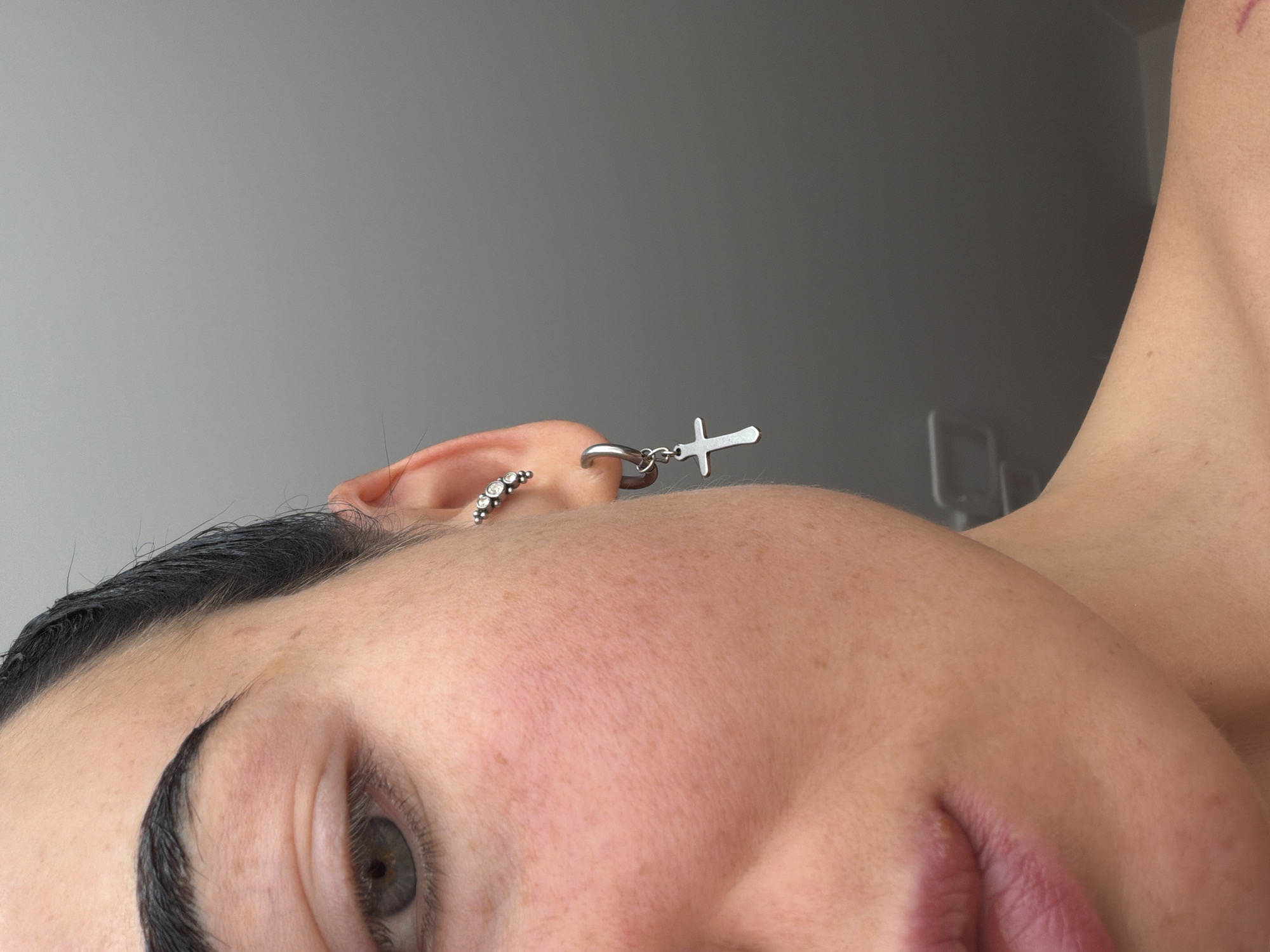 The image is a close-up, sideways portrait of a person, rotated 90 degrees counter-clockwise. The focus is on the right side of the person's face, including their eye, eyebrow, cheek, ear, and a portion of their neck and shoulder.
The person has a prominent, well-defined dark eyebrow and a visible blue or grey eye with dark eyelashes. Their skin appears clear with scattered light freckles on the cheek and forehead. Dark hair is visible along the top of the forehead. In the visible ear, there are two distinct earrings. One is a small hoop adorned with multiple clear, sparkling stones. The other is a larger silver-toned hoop from which a small, silver-colored cross-shaped dangle hangs. A sliver of the person's mouth, showing light-colored lips, is visible at the bottom of the frame.
The background is a plain, light grey wall, indicating an indoor setting. Faint outlines of what appears to be furniture or a fixture can be seen in the top right portion of the background. The lighting is soft and even. No specific activity is depicted other than posing for the photograph, and no particular emotion is discernible from the partial view of the face. There is no visible text in the image.