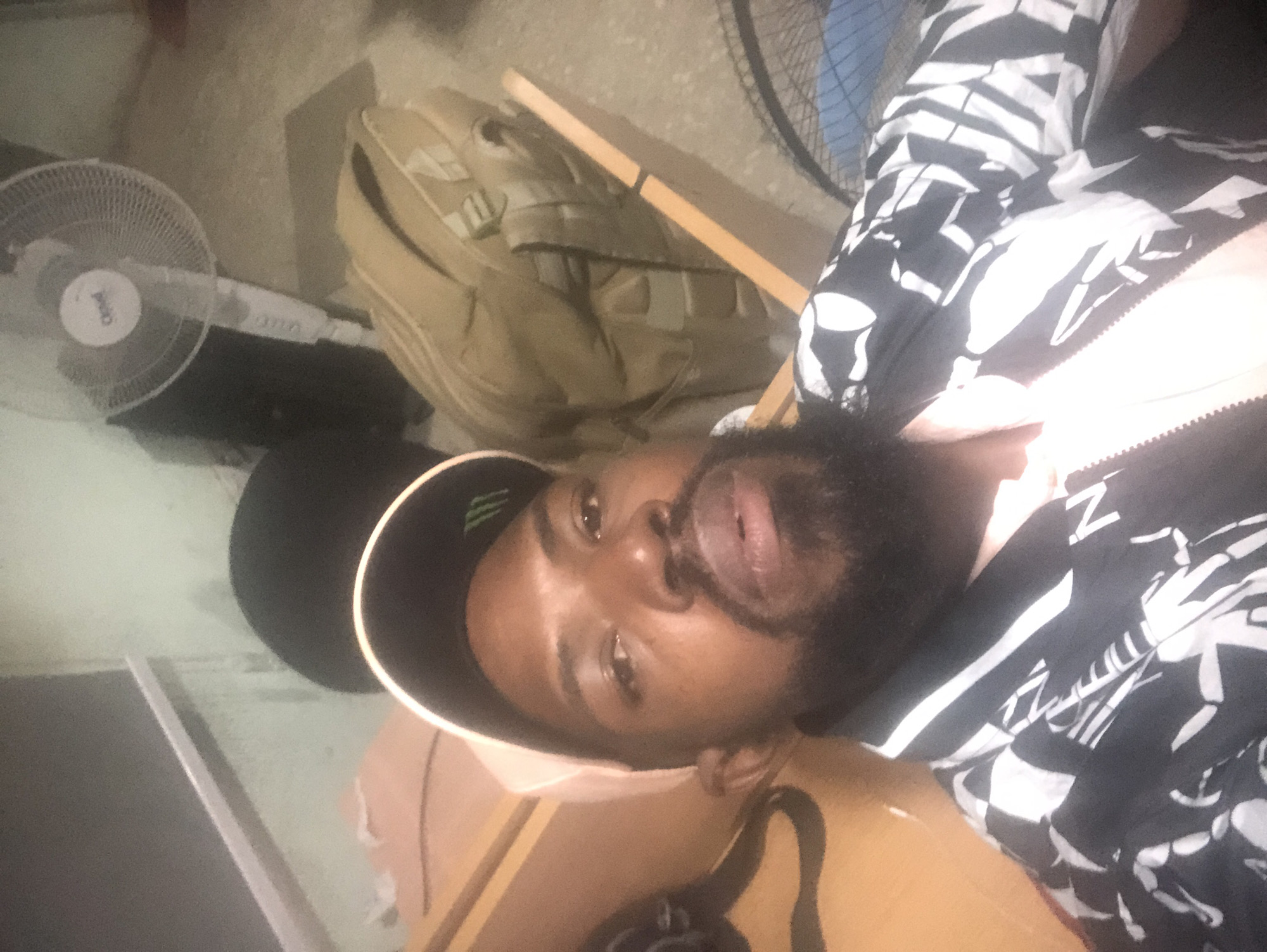 The image shows a medium shot of a Black man wearing a black baseball cap with a green logo, a light pink shirt, and a black and white patterned jacket. He has a beard and is looking at the camera. Behind him, there is a light green wall, a fan, a tan backpack, and wooden furniture. The scene appears to be indoors, possibly in a room or office in Ikeja, Nigeria.