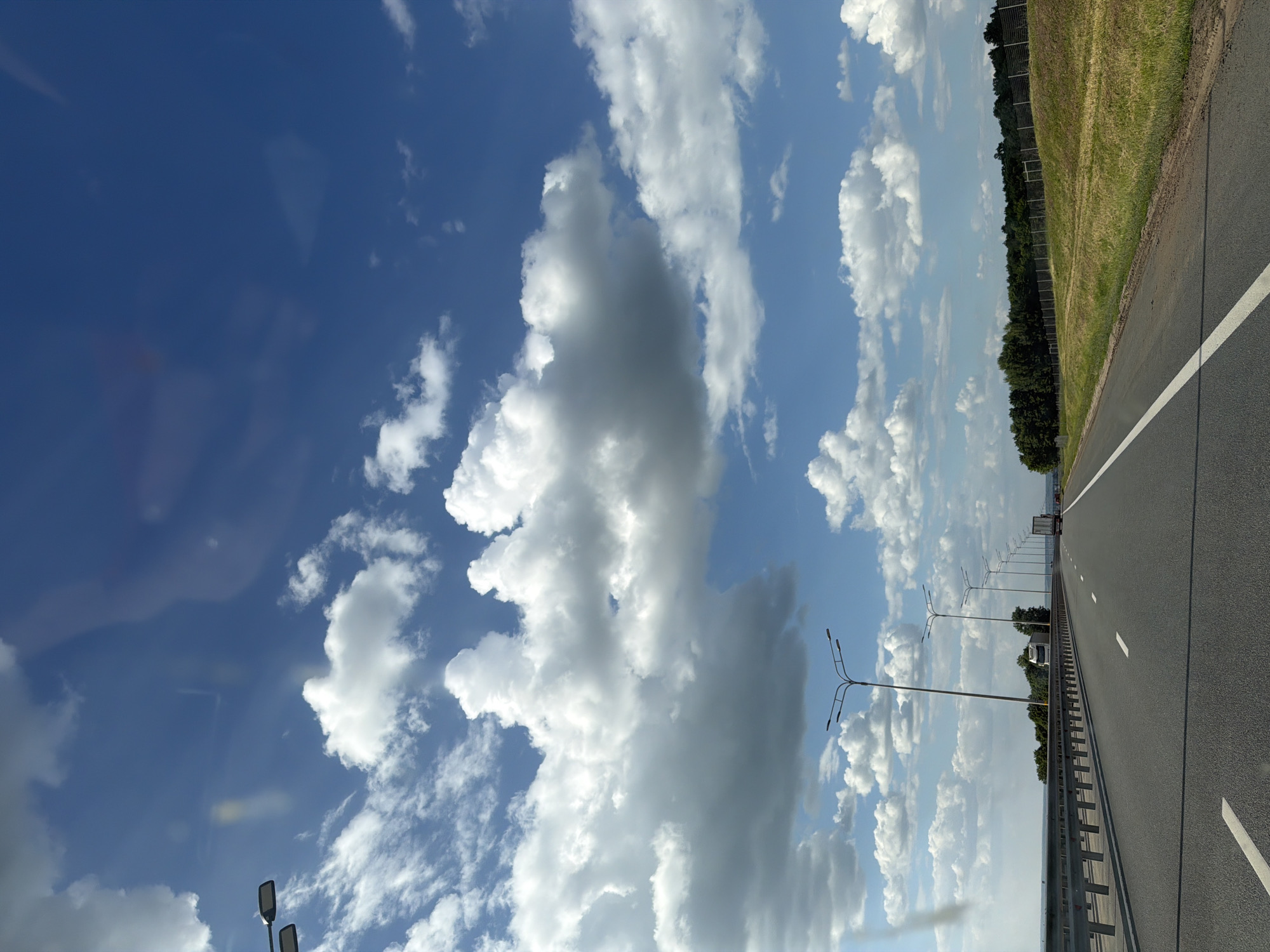 The image depicts a highway in the city of Shali, Russia, under a bright blue sky filled with fluffy white clouds. The perspective is from a vehicle moving along the road, looking forward. The asphalt road has clear white lane markings. On the left side of the road, there is a metal guardrail, and beyond that, a body of water or a wide expanse. Several lampposts line the left side of the highway. On the right side of the road, a grassy embankment slopes upwards, topped by a fence and a dense line of green trees. In the distance, a white truck is visible on the highway, and another vehicle can be seen further ahead. The scene is one of daytime travel on a sunny day.
