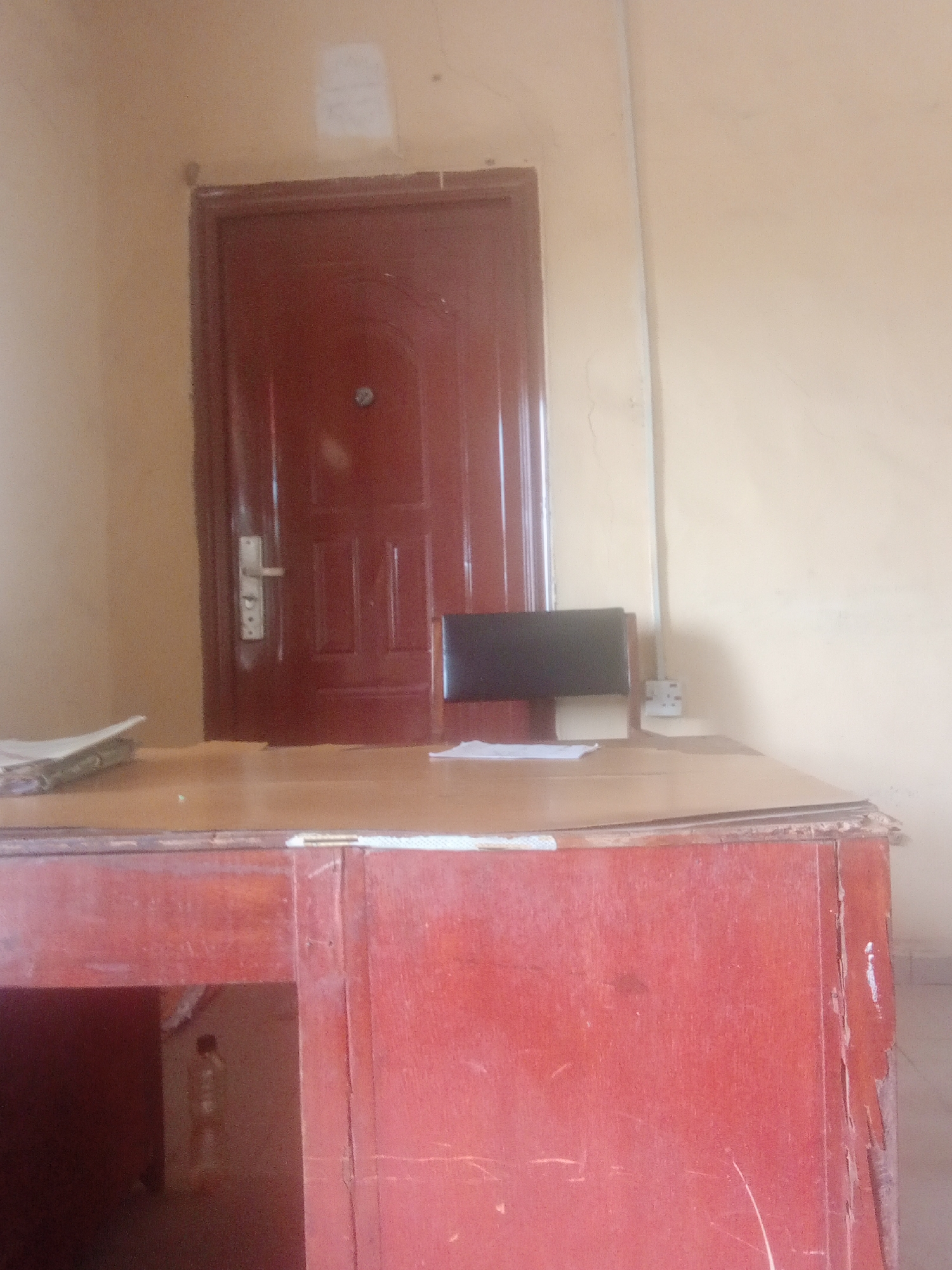 The image depicts an interior scene in Jalingo, Nigeria, focusing on a red wooden desk situated in front of a dark red door. The desk is positioned centrally in the frame, with its surface holding a stack of papers on the left and a black chair behind it. The door, which appears to be made of metal or reinforced wood, is closed and has a silver handle. To the right of the door, a white electrical outlet is visible on the wall. The walls are painted a pale yellow and show signs of minor wear. The floor is not visible. The overall impression is one of a simple, utilitarian office or room.