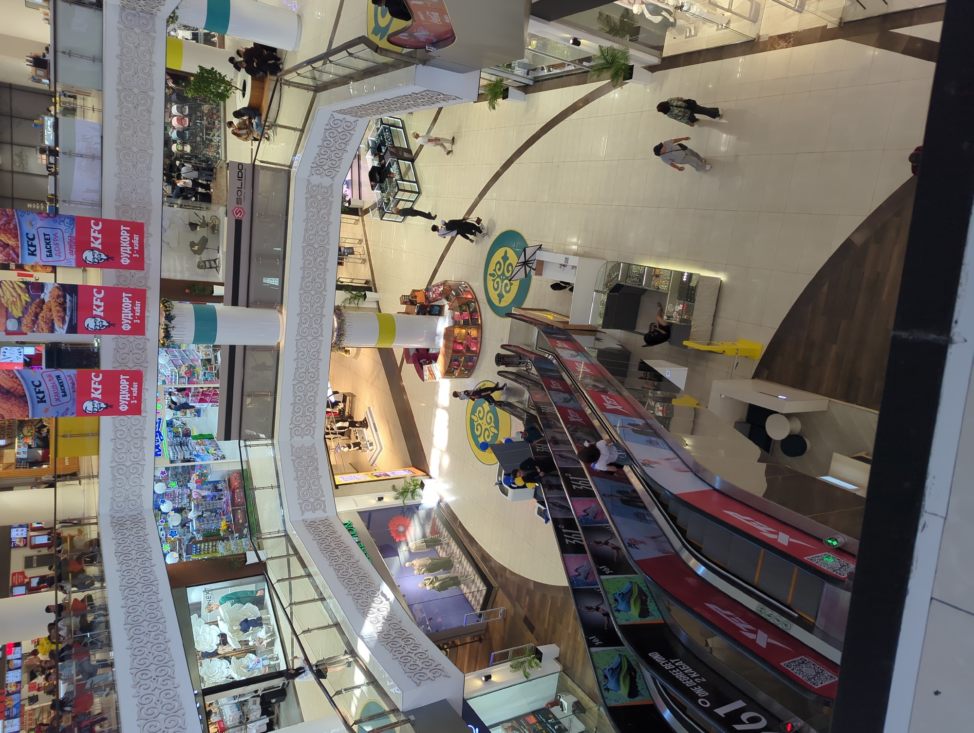 This is an indoor scene taken from an elevated perspective, likely within a shopping mall located in Bishkek City, Kyrgyzstan. The multi-level mall features various retail stores and food establishments. In the foreground, escalators carry people between floors. Prominent signage for KFC is visible, advertising "food court 3 floor."
The mall's architecture includes white pillars with decorative patterns along the railings. People are seen walking through the mall's corridors. The mall includes retail stores, and there are seating areas with tables and chairs, where people are sitting.