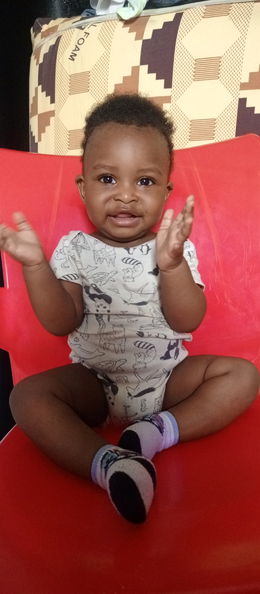 A dark-skinned baby with dark, curly hair is seated on a bright red chair, looking directly at the viewer with wide, dark eyes and an open-mouthed smile. The baby's hands are raised to chest height, palms open, in a gesture that suggests clapping or waving.
The baby is wearing a light-colored, possibly white or cream, short-sleeved bodysuit adorned with various black and white animal illustrations, including raccoons, turtles, cats, giraffes, pandas, llamas, and also an anchor, a whale, and a fish. On the baby's feet are socks with black and white patterns, complemented by light blue and purple bands around the ankles.
Behind the baby, primarily visible above their head, is a patterned cushion or mattress. This object features a geometric design in shades of brown and beige. The word "FOAM" is distinctly visible on this cushion, oriented vertically along one edge. The setting appears to be indoors, simple and uncluttered. The baby conveys a sense of happiness and engagement.