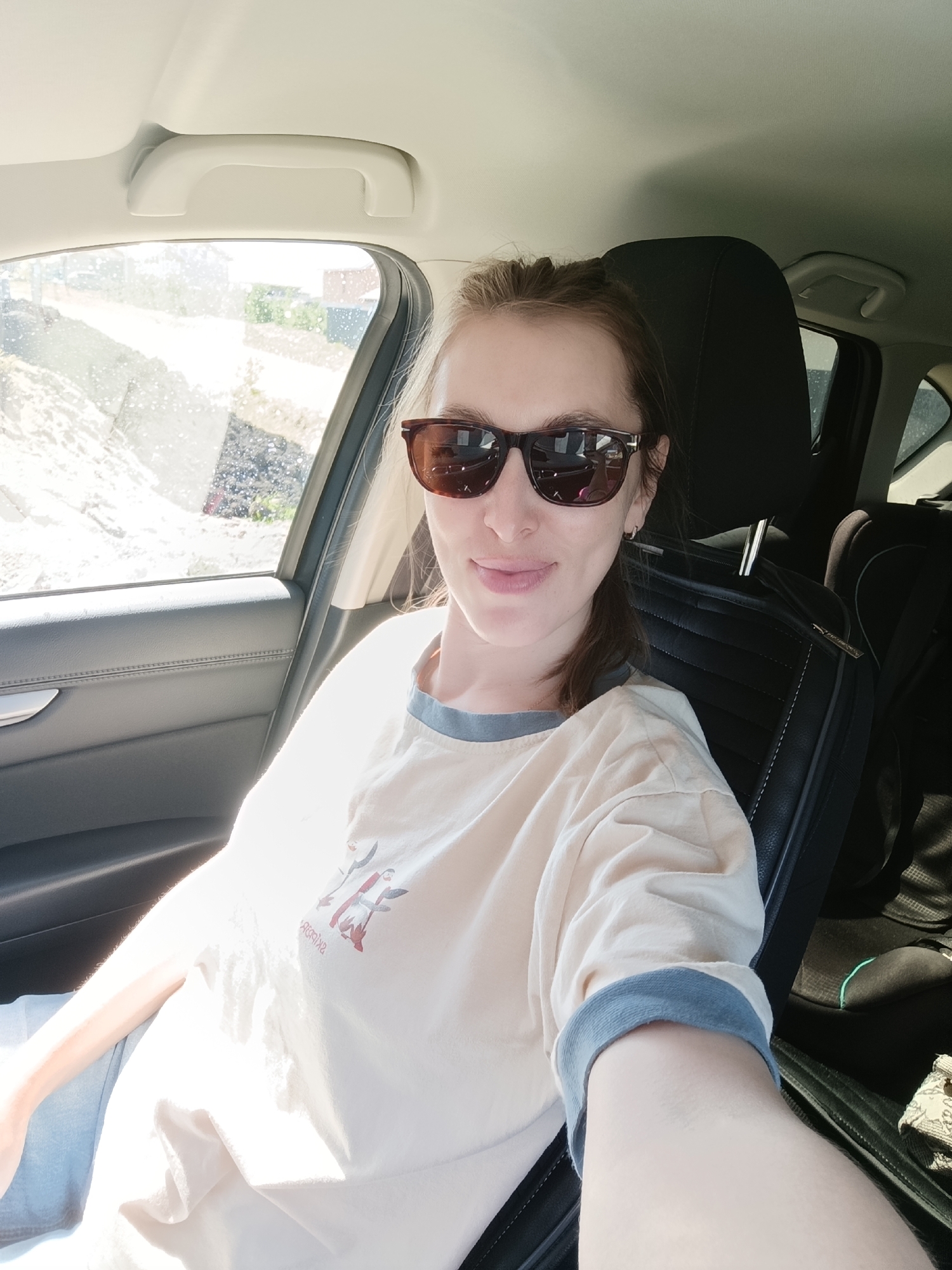 A woman is sitting in the driver's seat of a car. She is wearing sunglasses and a light-colored t-shirt with blue trim. The car is parked next to a construction site. The location is the city of Chaikovsky, Russia.