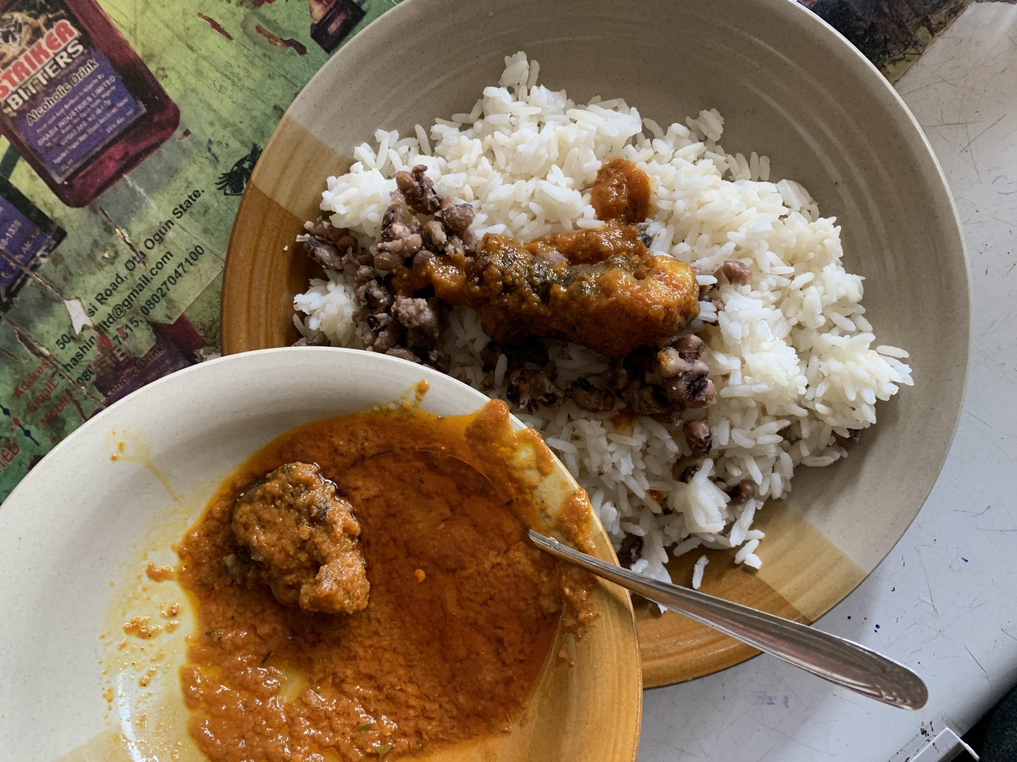 The image shows two plates of food. The larger plate contains white rice mixed with beans and topped with a reddish-brown sauce and a piece of fried meat. The smaller plate contains a reddish-brown sauce with a small chunk of meat in it and a spoon resting on the plate. The plates are placed on a surface with a printed background that mentions "Striker Bitters Alcoholic Drink" and "Ota Ogun State", Nigeria, but it does not have information about Kabong.