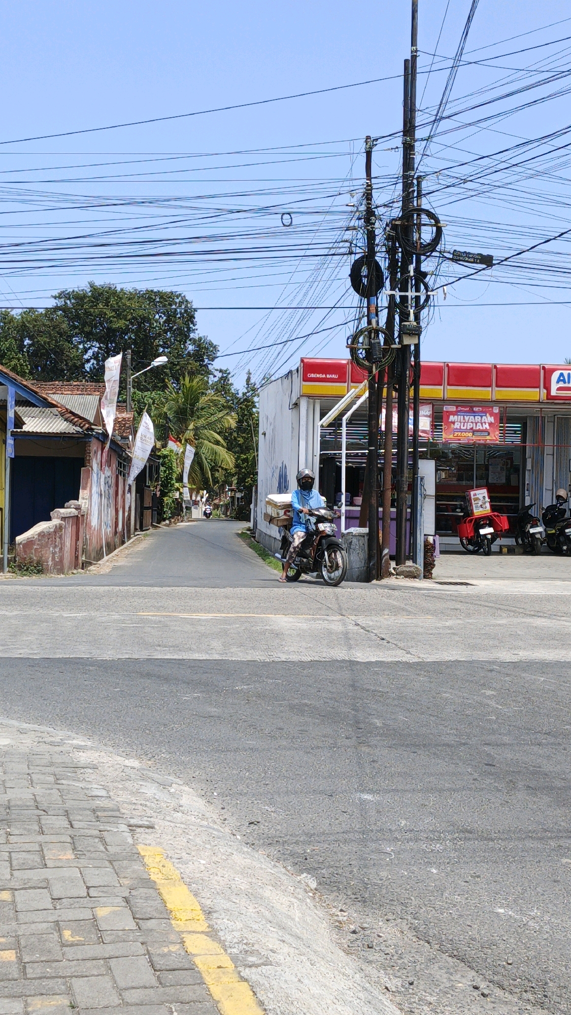 A medium shot captures a street scene in Cibenda, Indonesia. The sun is shining brightly, creating clear shadows. A person wearing a blue shirt, dark pants, a helmet, and a face mask rides a motorcycle. The motorcycle is loaded with several cardboard boxes, possibly merchandise, strapped to the rear. The rider is positioned in the center of the frame, stopped at an intersection.
To the right of the motorcycle is a building with a red and yellow awning that appears to be a convenience store or gas station, indicated by the signage with "ALFA" visible. There are other motorcycles parked in front of this building. A utility pole with numerous wires is prominent on the right side of the image, adjacent to the store.
To the left of the motorcycle and road, there are buildings with a more traditional Indonesian architectural style, featuring tiled roofs and colorful facades, some adorned with small flags. Palm trees are visible in the background, suggesting a tropical environment. The road itself is paved, with a yellow line marking the edge of a pedestrian walkway or bicycle lane made of interlocking concrete pavers in the foreground. The overall impression is a typical, bustling street in a populated area of Indonesia.