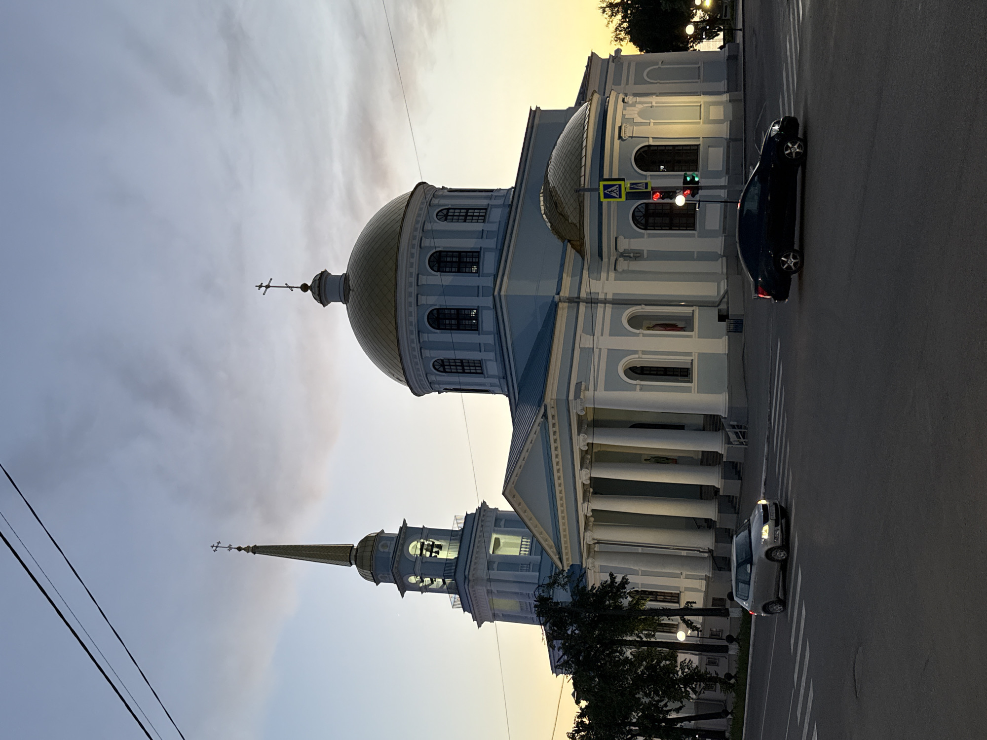This media file captures a street scene in Votkinsk, Russia, featuring a prominent cathedral or church during dusk or early evening. The central subject is a large, stately religious building, predominantly light blue and white, adorned with classical architectural elements such as columns, arched windows, and cornices. It features a large, golden, onion-shaped dome topped with an Orthodox cross on its main structure, and a tall, slender bell tower or spire to its left, also crowned with a golden top and an Orthodox cross. Lights illuminate the interior of the bell tower.
The sky above displays a blend of soft grey clouds and a warm, yellowish-orange glow, indicative of either sunset or sunrise, contributing to the tranquil atmosphere. Streetlights and lights on the cathedral facade are illuminated, casting a gentle glow on the building and its surroundings.
In the foreground, an asphalt road runs horizontally across the frame. A dark-colored sedan is visible on the right side of the road, positioned near a pedestrian crossing sign and a traffic light displaying both red and green signals. A lighter-colored car is partially visible further down the road to the left. Utility lines are stretched across the upper left portion of the frame. The overall scene is peaceful, highlighting the architectural beauty of the cathedral against the twilight sky.