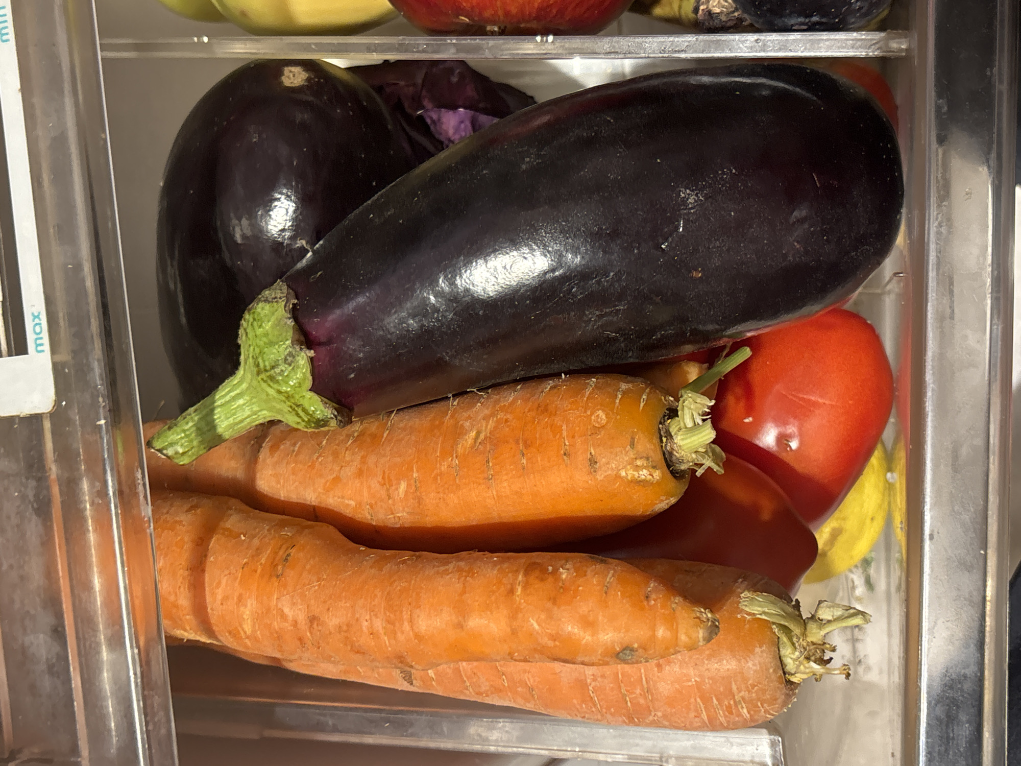 In the image, a clear drawer holds an assortment of fresh vegetables. Two long, orange carrots with green leafy tops rest at the bottom. Above them lie two large, dark purple eggplants, one rounder and one elongated. A red tomato is nestled among the carrots and eggplants. A piece of red cabbage is also visible. Above, glimpses of yellow and red fruits or vegetables are visible. The drawer appears to be inside a refrigerator. The setting is likely a residential kitchen. There is no information to pinpoint this to Buenos Aires, Argentina, or any other specific location.