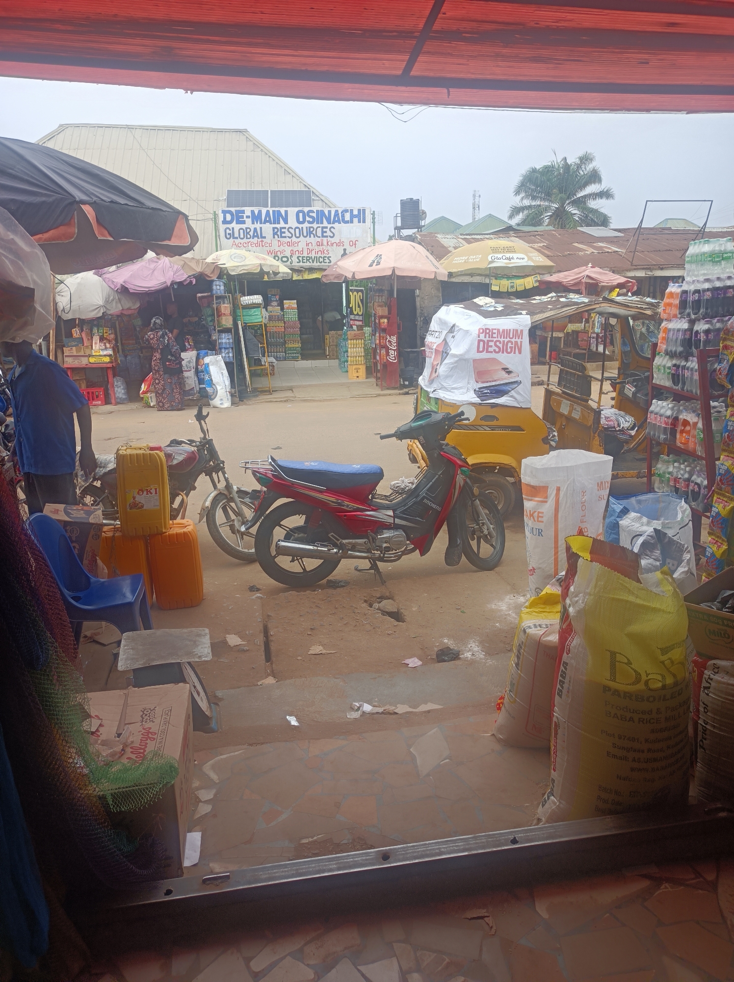 The image shows a view from inside a shop in Narayi, Nigeria, looking out onto a busy street scene. In the foreground are bags of rice, and a box of Kellogg's cereal. To the left is a man standing, dressed in blue. In the middle ground are two motorcycles parked near the edge of the shop. One motorcycle is red and the other is brown. Also in the middle ground are several yellow jerrycans, a blue plastic chair, and a small grey stool. In the background, there is a building with a sign that reads "DE-MAIN OSINACHI GLOBAL RESOURCES." The street is filled with people, other shops, and several yellow "Keke Napep" auto-rickshaws. Several umbrellas provide shade to the shops and pedestrians.