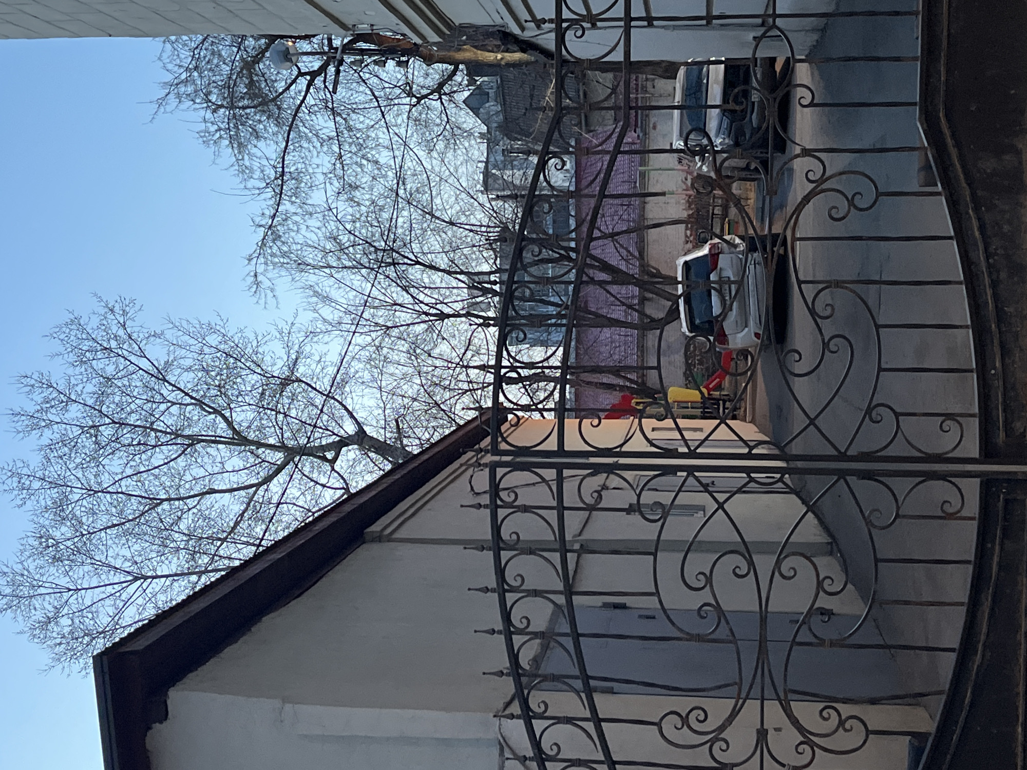 The image captures an outdoor residential scene, framed by a decorative wrought iron gate on the right side. On the left, a white building with a dark brown gabled roof is partially visible, its facade showing some signs of wear.
Bare tree branches, devoid of leaves, stretch across the upper and central parts of the frame, silhouetted against a clear, bright blue sky. These trees suggest either winter, late autumn, or early spring. A light fixture or security camera is attached to a branch in the upper right.
Through the intricate scrollwork of the gate, a paved area or driveway in the background reveals two parked vehicles: a white SUV and a darker car. Behind these vehicles, a low, pinkish-red wall is visible, with glimpses of other structures beyond. A small, colorful object, possibly children's play equipment in yellow and red, can be seen near the parked cars.
The scene is brightly lit by natural daylight, indicating it is daytime. No people are present, and no specific activity is occurring. There is no visible text in the image. The overall impression is that of a quiet residential area during a cooler season.