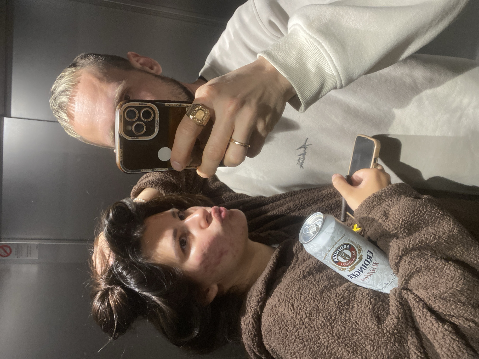 A reflection of two people taking a selfie in what appears to be an elevator. The man is holding a cell phone, with a gold ring on his ring finger and a larger, decorative ring on his middle finger. He's wearing a white sweatshirt. The woman in the reflection is holding a can of Erdinger beer, wearing a brown fuzzy robe, and has her hair up. The elevator's wall is a gray/silver color. The location in the city of Kiryat Bialik, Israel, cannot be verified from the image.