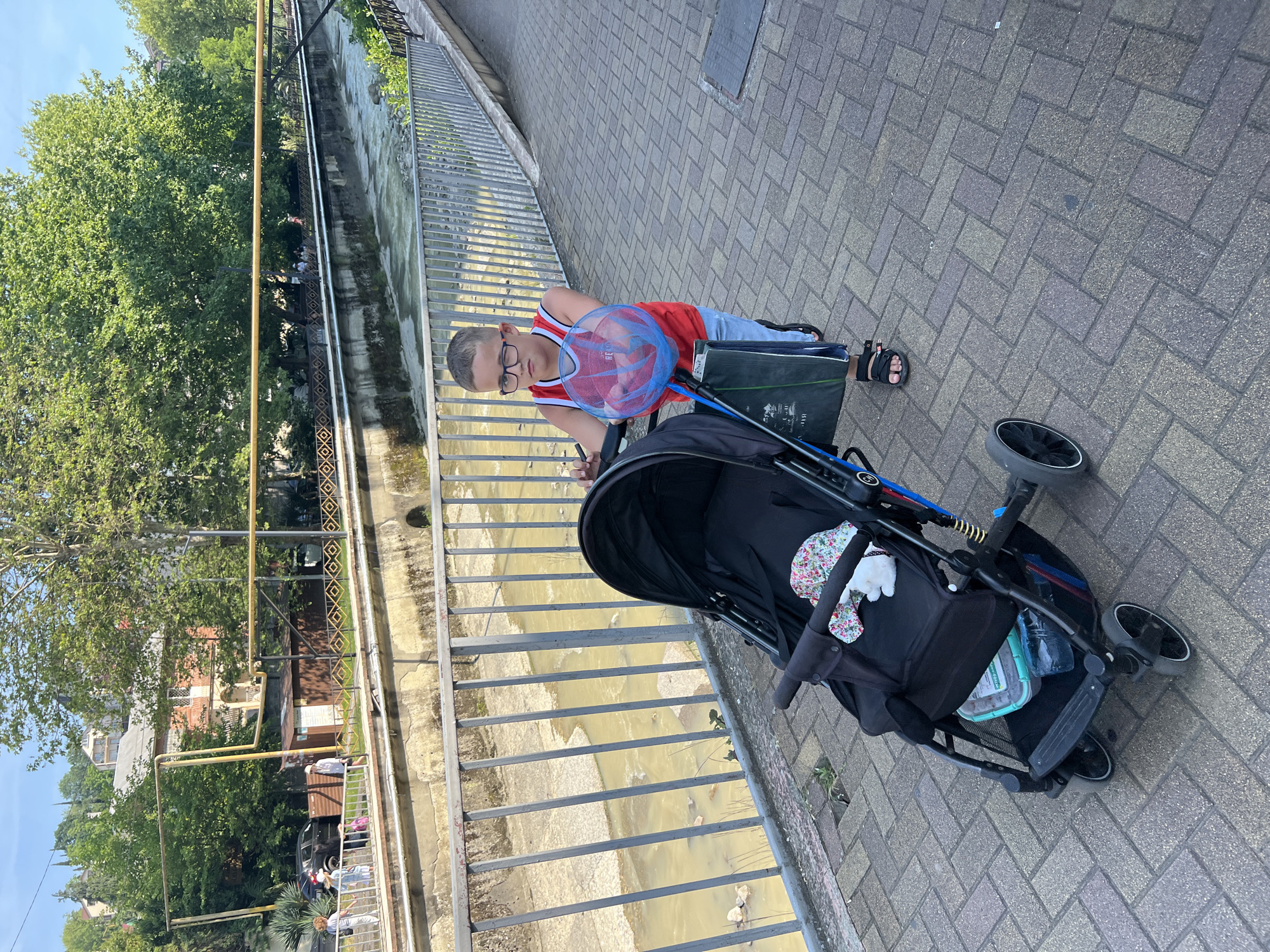 A young boy, appearing to be in his early teens, is positioned on a paved pedestrian walkway, pushing a black baby stroller. He is wearing black-rimmed glasses, a red and white sleeveless basketball jersey, light blue shorts, and black sandals. His hair is short and light-colored. He holds the stroller's handle with both hands. A red and blue basketball contained within a net hangs from the stroller's left side. A dark green reusable bag is also suspended from the stroller frame. Inside the stroller's seat, a white plush toy wearing a floral pattern hat is visible. Beneath the stroller's seat, a teal plastic container with a lid and a plastic water bottle are stored.
The walkway is paved with rectangular grey and brown interlocking pavers arranged in a herringbone pattern. To the boy's left, a grey metal railing with vertical bars separates the walkway from a shallow, murky yellowish-brown river or canal. The waterway features concrete banks, and some rocks are visible in the water.
In the background, across the river, dense green trees and foliage are prominent under a clear blue sky. Visible through the trees on the left side are a multi-story building with reddish-orange walls and white trim, featuring balconies and a pitched roof, as well as yellow industrial pipes running horizontally and vertically. A small dark-colored vehicle and a brown utility shack are also discernible. Further along the opposite bank, a decorative metal fence with diamond patterns is present. Several indistinct figures can be seen walking in the background on the far side of the river. The scene is brightly lit by natural sunlight, indicating daytime.
This scene is located in the city of Sochi, Russia.