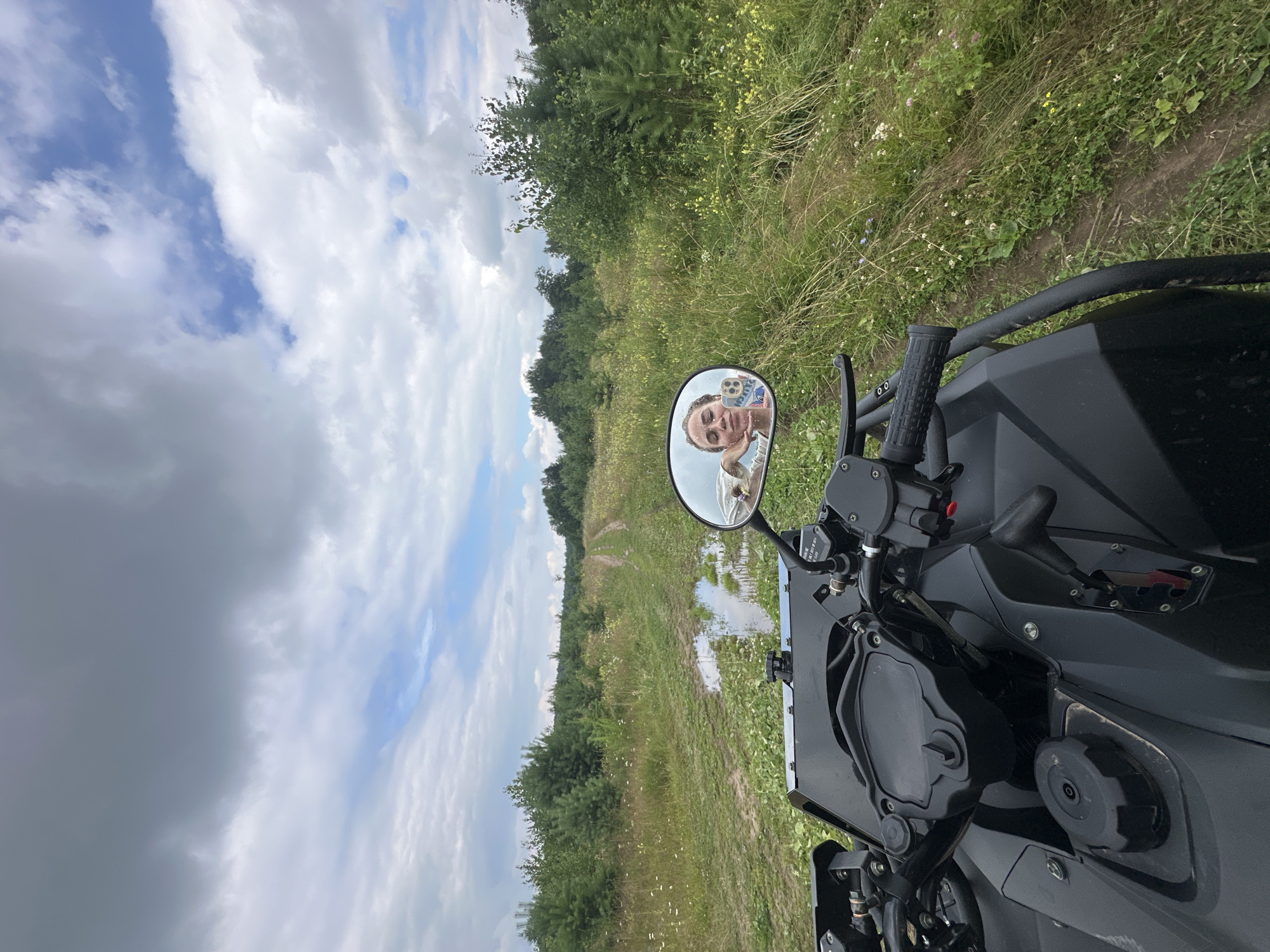 The image shows a black ATV parked on a dirt path in a rural area. The path leads through a grassy field with trees on either side. The sky is partly cloudy. Reflected in the ATV's side mirror is a young woman taking a selfie. The location is identified as Kharnavy, Russia.