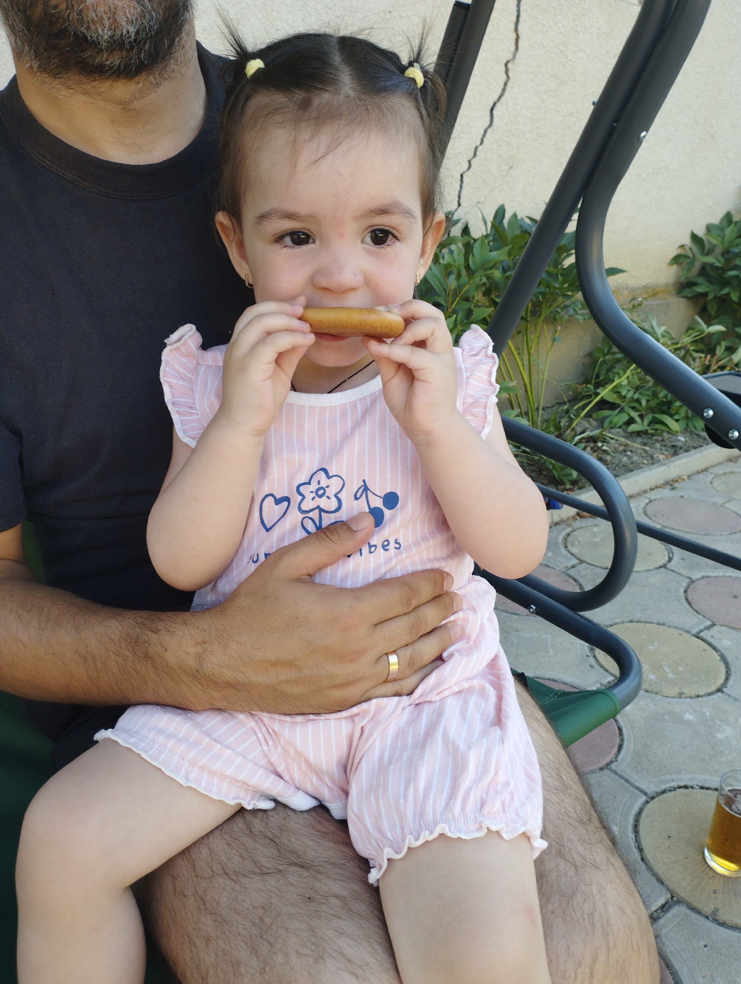 A young child with dark hair in pigtails, wearing a pink and white striped outfit, sits on the lap of a bearded adult wearing a dark t-shirt. The child is holding a small, elongated food item to their mouth with both hands. The adult's left arm is around the child, and a gold ring is visible on the adult's finger. They are seated on a green chair, part of a swing set, outdoors. In the background, there is a light-colored wall, green foliage, and a paved ground with circular patterns. A glass of amber liquid is visible in the lower right corner. The scene appears to be in the city of Fălești, Moldova.