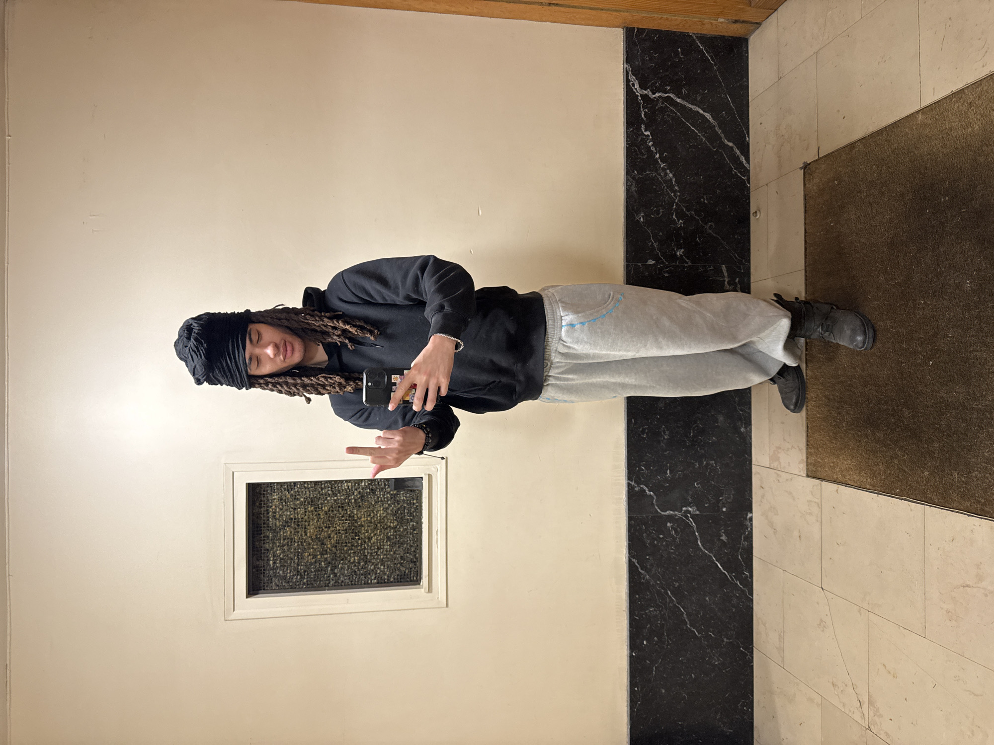 The scene depicts a single individual standing centrally within an indoor hallway or vestibule in an unspecified building in the city of Paris, France. The individual, appearing to be a young adult, has long dreadlocks and wears a black head covering. They are dressed in a black hooded sweatshirt and light grey sweatpants with a light blue accent stripe around the left pocket. Dark boots are worn on their feet. A silver-colored chain bracelet is visible on their left wrist.
The individual holds a black smartphone with a patterned case in their right hand, positioned near their chest. Their left hand is raised, with the middle finger extended upwards, directed towards the viewer or camera. Their facial expression is neutral, with eyes partially closed or cast downwards.
The environment features light beige or off-white painted walls. A dark, possibly black, marble wainscoting with prominent white veining runs horizontally along the lower third of the walls. To the left, a rectangular window or opening with a light frame contains a textured or gridded glass pane. The floor is covered with light-colored square tiles, and a dark brown, rectangular doormat or rug is situated on the floor in the foreground, partially beneath the individual's feet. A wooden doorframe or panel is visible on the far right.