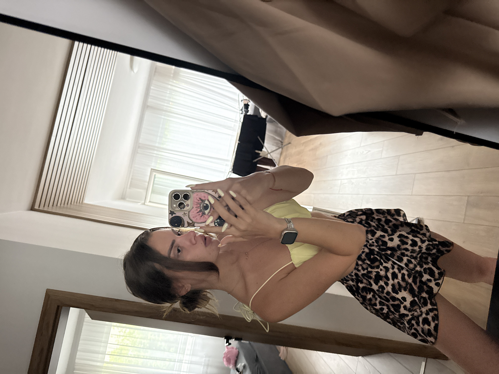 A young woman is taking a mirror selfie in what appears to be a bedroom. She is wearing a yellow crop top with spaghetti straps and a leopard print skirt. Her hair is pulled back loosely, and she is holding a phone with a decorative case. The room has light wood flooring, white sheer curtains, and a doorframe visible on the left. A portion of a tan garment is draped in the foreground on the right side of the image. The reflection in the mirror shows a white ceiling with horizontal wooden slats, and further into the room, a table covered with a black cloth. The scene does not contain any elements that would indicate it is located in the city of Plovdiv, Bulgaria.
