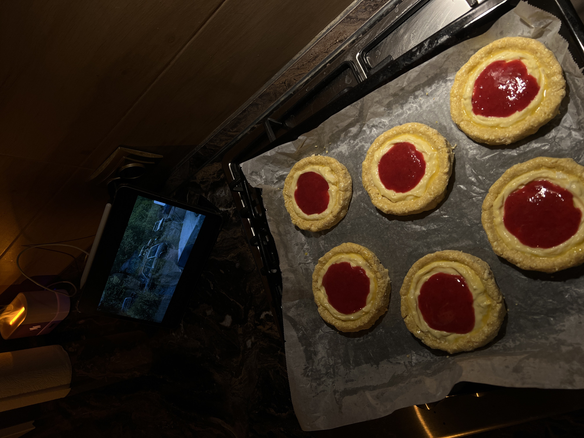 The image depicts a close-up, high-angle shot of six Danish pastries arranged on a baking sheet lined with parchment paper. The pastries are circular with a golden-brown crust, a creamy white filling, and a vibrant red jam center. The baking sheet is placed on a dark, speckled countertop, possibly marble, with a metallic oven rack visible to the right. In the background, a tablet computer is propped up, displaying an image of several vehicles in a wooded area. To the left of the tablet, a white paper towel roll and a small, illuminated humidifier or diffuser are present. The overall lighting suggests an indoor setting, possibly a kitchen, with warm, ambient light. There is no indication of the city of Ternopil, Ukraine, in the image.
