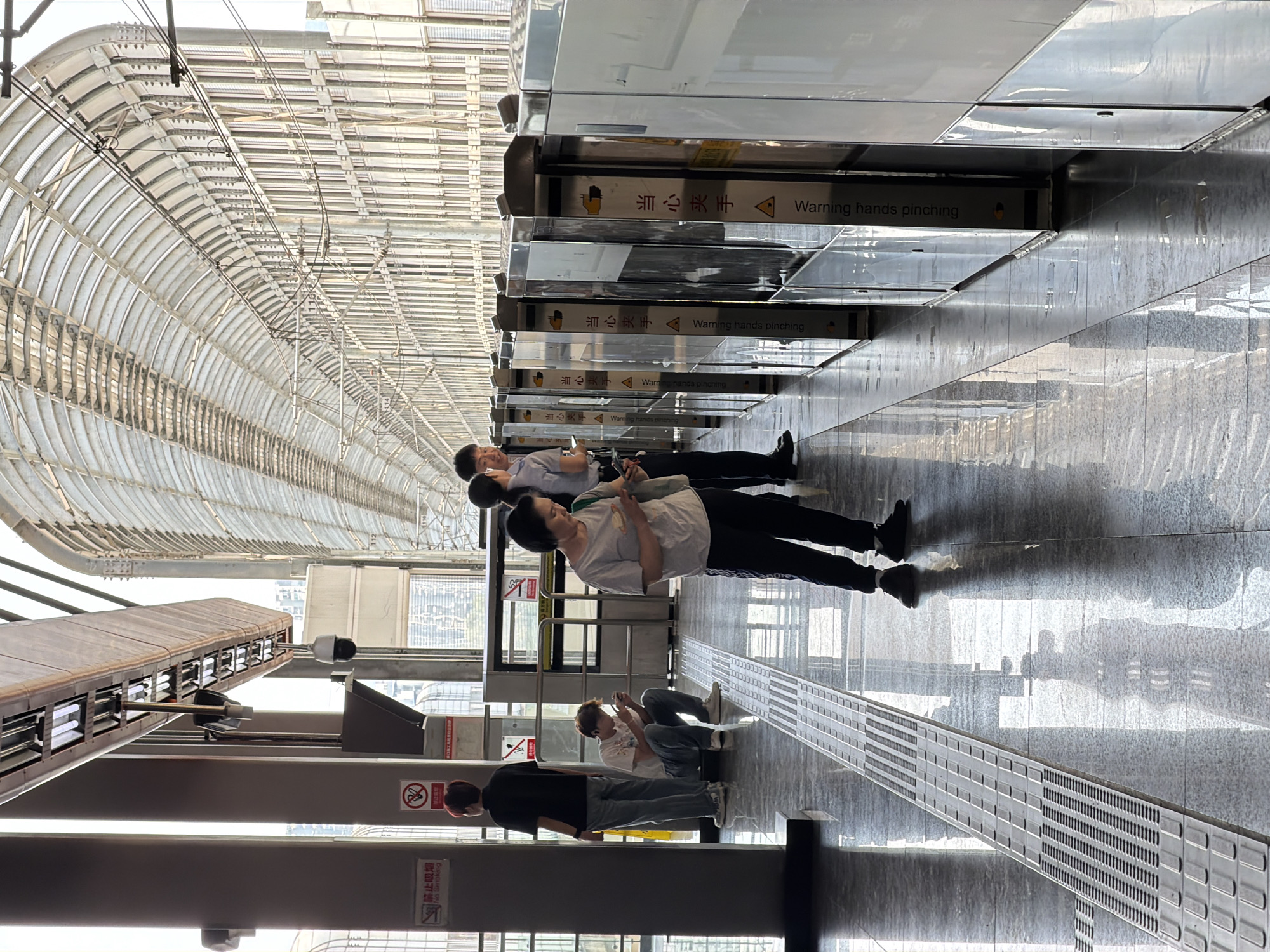 The image depicts a covered metro station platform in Tiaodeng, China. The platform is characterized by a polished, reflective floor and a long, transparent roof structure supported by a metal framework. A row of automatic doors, some open and some closed, line the edge of the platform, leading to what appears to be the train.
Several individuals are present on the platform. In the foreground, a woman in a grey t-shirt and dark pants is looking at her phone. To her left, a man in a black t-shirt and blue jeans stands looking forward. Another man, wearing a white t-shirt and blue jeans, is crouched down, also looking at his phone. Further back, two other individuals are partially visible near the doors. There are signs visible on the platform pillars, including a "No Smoking" sign. The overall atmosphere suggests a typical scene in a busy urban transit hub.
