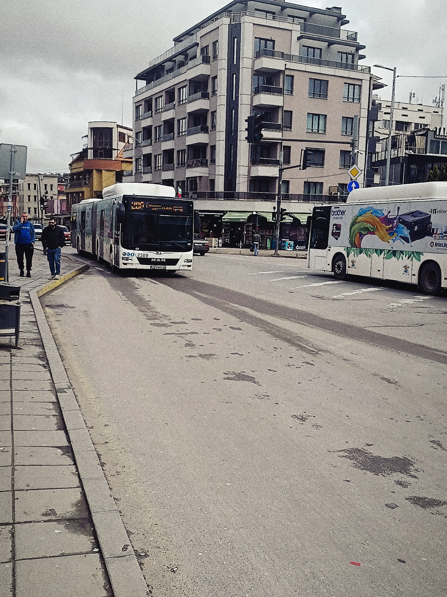 The image depicts a street scene in Sofia, Bulgaria, on an overcast day. The foreground features a section of a sidewalk paved with square concrete slabs, adjacent to a wide asphalt road showing dark marks indicating recent rain or cleaning. On the sidewalk, two men walk side-by-side; the first man wears a blue jacket and speaks on a mobile phone, while the second man is dressed in darker clothing.
In the roadway, a white, articulated MAN city bus with the license plate CB 4945 HM is moving towards the camera, its destination display reading in Cyrillic script. To the right, another bus, partly out of frame, is adorned with a colorful advertisement for Brother printers. Buildings of varying heights and architectural styles form the backdrop, with a prominent multi-story building in beige and gray colors with balconies and a top floor apartment visible. Traffic lights are mounted on poles at the crosswalk. Street signs, a traffic island, and other street furniture are sparsely placed in the scene. A store called "SHISHAA" is visible in the middle-ground with green awnings.