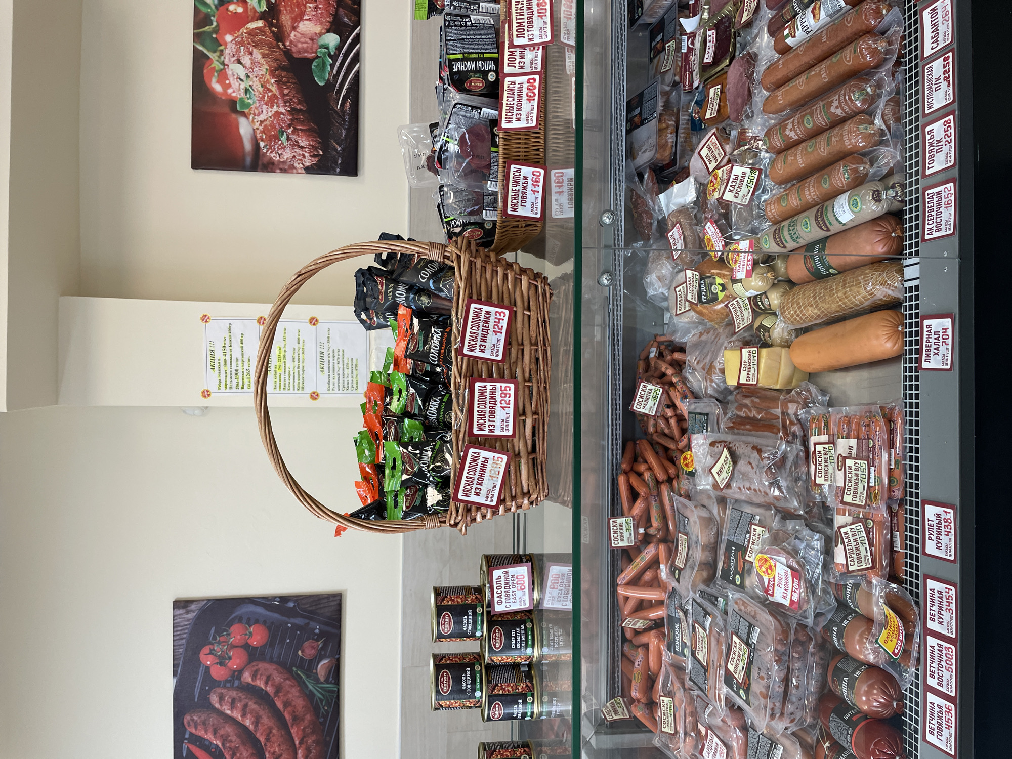 The image shows the interior of a meat shop or a deli counter within a larger store, likely in Almaty, Kazakhstan, given the Cyrillic text on the product labels. The focus is on a display case filled with various types of sausages, cured meats, and canned goods.
In the foreground, a refrigerated glass display case showcases rows of packaged sausages, including "сосиски" (sausages) of different varieties like "конские" (horse meat) and "говяжьи" (beef). There are also some bulkier meat products and possibly cheese. Pricing labels are visible for many of these items.
Above the display case, a large woven basket overflows with packaged meat products, likely jerky or dried sausages, with prominent Cyrillic labels such as "мясная соломка" (meat straw) and "чипсы мясные" (meat chips). Several cans are also stacked to the left of the basket.
The background features two large posters on the wall. The poster on the left depicts grilled sausages with tomatoes and herbs, while the poster on the right shows a cooked steak with herbs. Various promotional signs and small circular stickers with promotional text are also attached to the wall and the display.
The overall environment appears to be a retail setting with bright overhead lighting, creating a clean and well-lit display of food products. No people are visible in the image. The detailed labeling suggests a local market environment where consumers can purchase a variety of meat products.