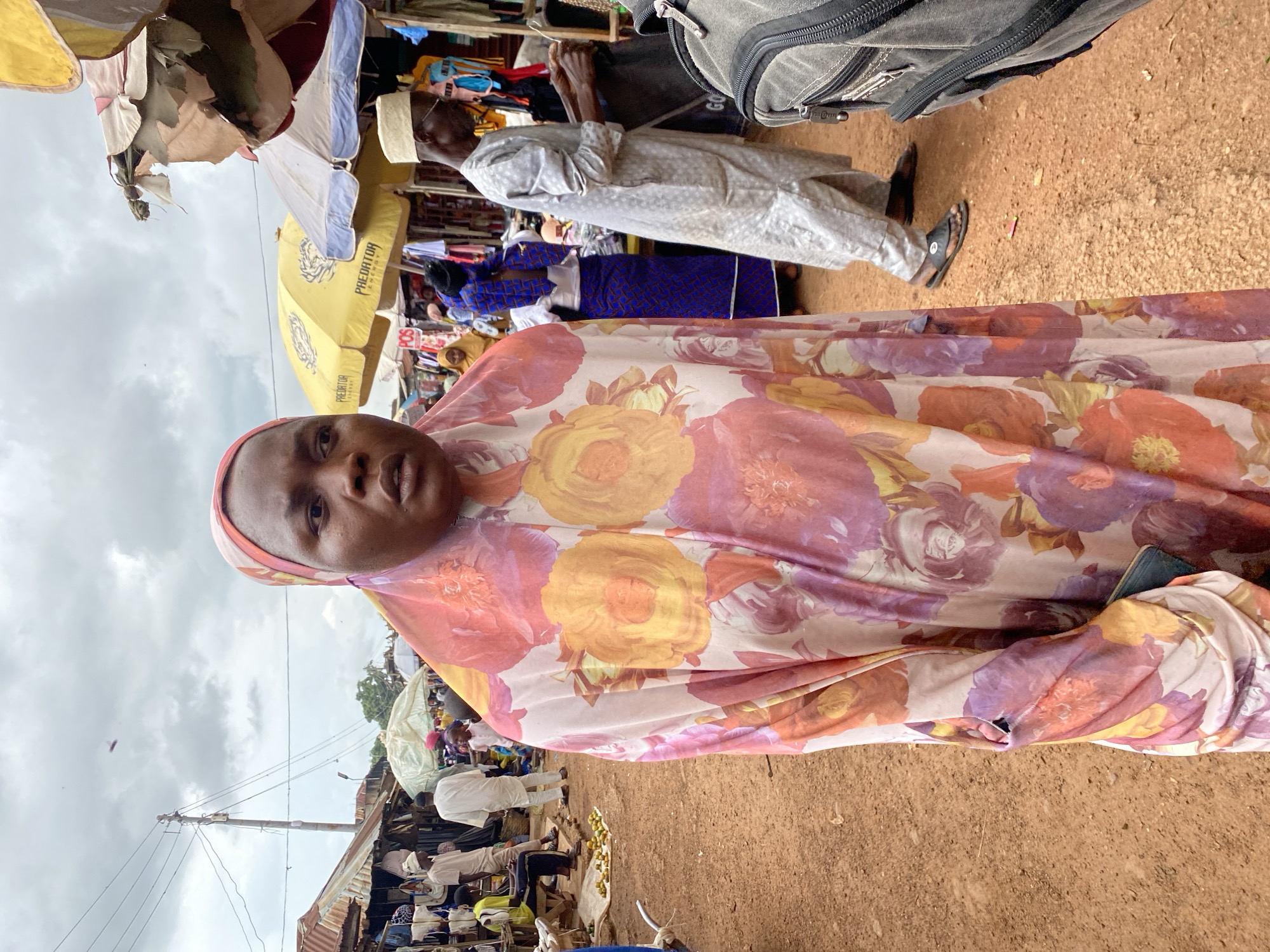 The image shows a marketplace scene in Bukuru, Nigeria. The foreground is dominated by a person wearing a vibrant, floral-patterned headscarf and dress. Behind them, the market unfolds with various stalls and people. To the left, umbrellas are set up, some appearing worn and torn, providing shade. One yellow umbrella has the text "PREDATOR ENERGY" visible. In the background, several individuals are seen, some in traditional attire. One man is wearing a light-colored, patterned robe, and another person in the mid-ground is dressed in a blue and patterned outfit. The ground appears to be dirt, common in outdoor market settings. The sky is overcast, suggesting a cloudy day. The overall impression is of a bustling, everyday market in a Nigerian community.
