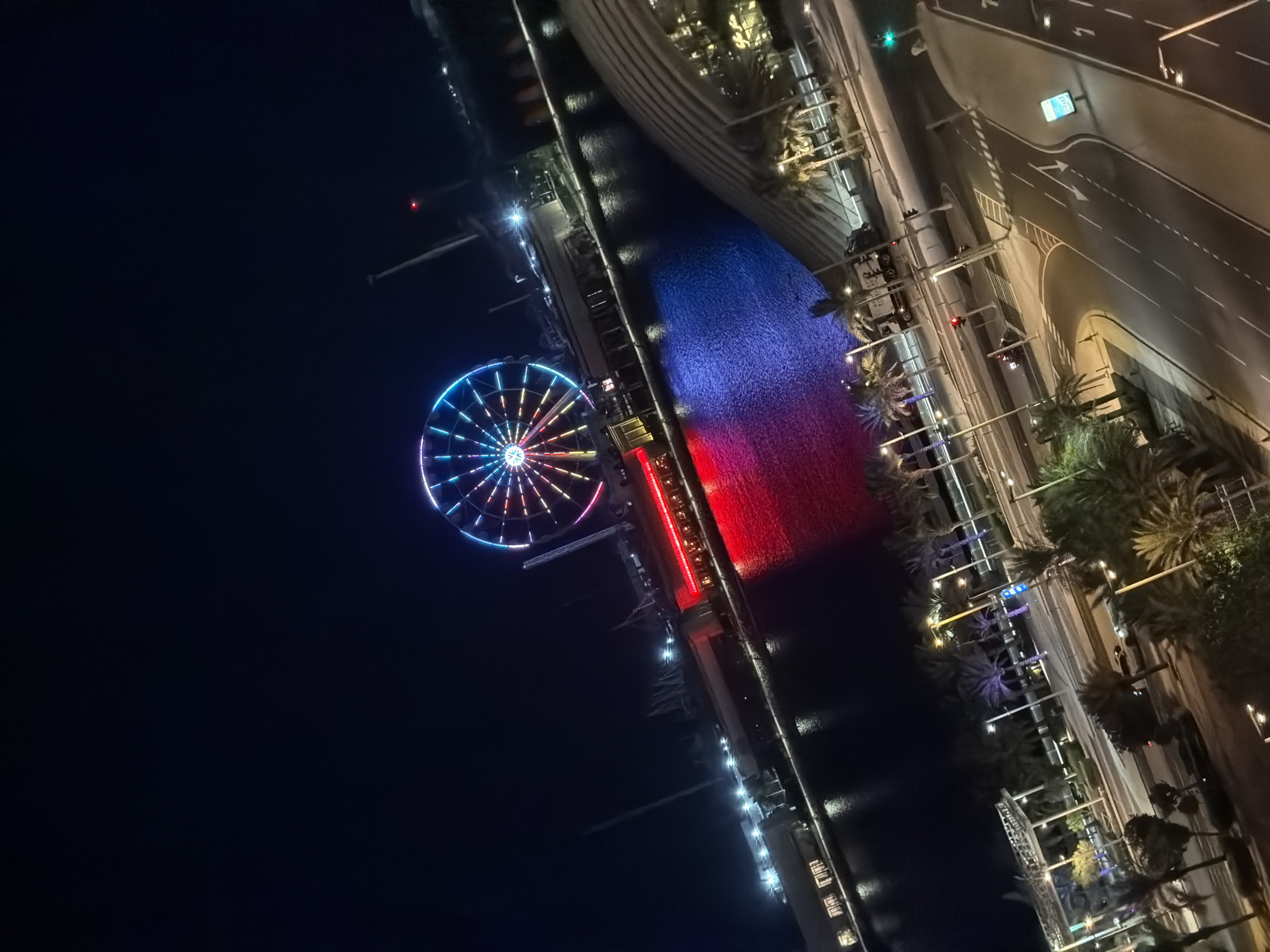 An aerial, angled, nighttime shot shows a Ferris wheel with multicolored lights dominating the upper half of the frame. The Ferris wheel is situated on a waterfront. A building with red lights along its facade is visible below the Ferris wheel. The water in front of this building is illuminated by red and blue lights. In the foreground, a street scene with palm trees, streetlights, traffic lights, and a road is depicted. Vehicles, including a truck and at least one motorcycle, are present on the road. The overall scene suggests an urban entertainment district at night.