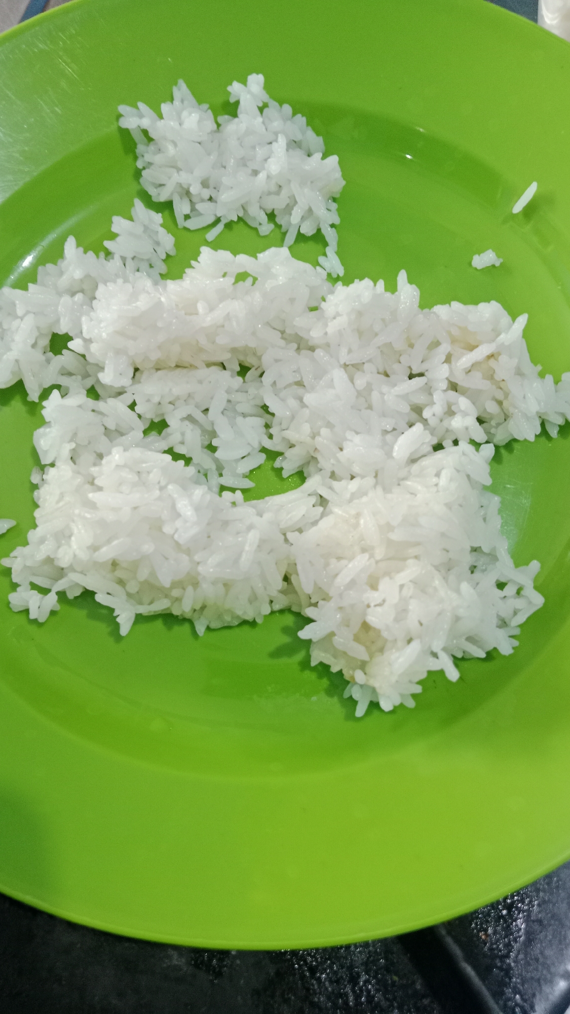 The image shows a close-up of a bright green plastic plate containing a pile of white rice. The rice is arranged in several distinct clusters, with some grains scattered across the plate. The plate is resting on a dark, possibly black, textured surface. The lighting suggests an indoor setting, likely a kitchen or dining area. There are no people or other objects visible in the frame, and the focus is solely on the rice and the plate. The image does not provide any specific visual cues that would allow for identification of its location in Denpasar, Indonesia.