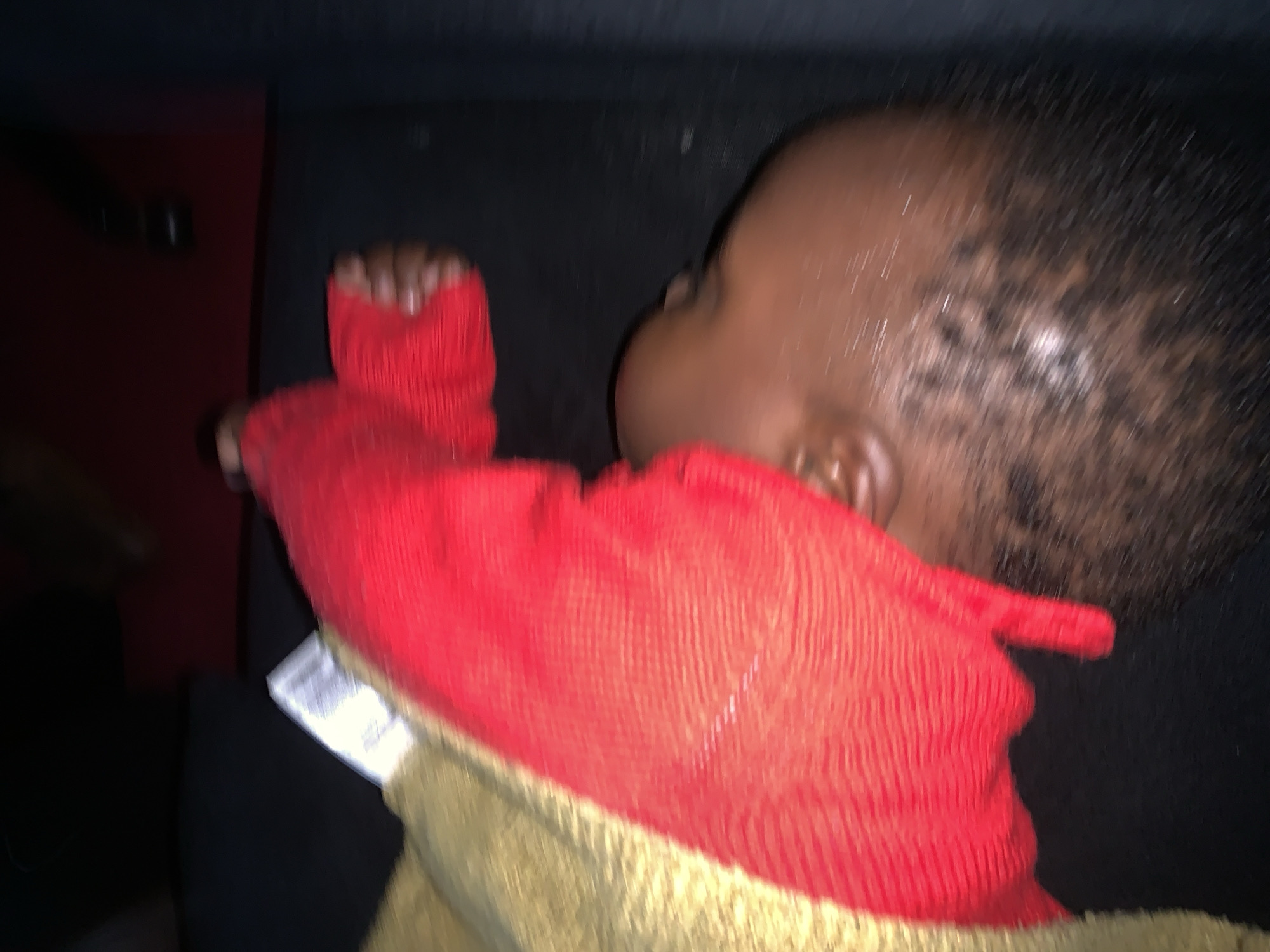 A close-up, blurry, low-angle shot shows the head of a dark-skinned person lying down. The person is wearing a red textured garment over their shoulder and arm, and a tan-colored fabric is visible on the left. The background is dark.