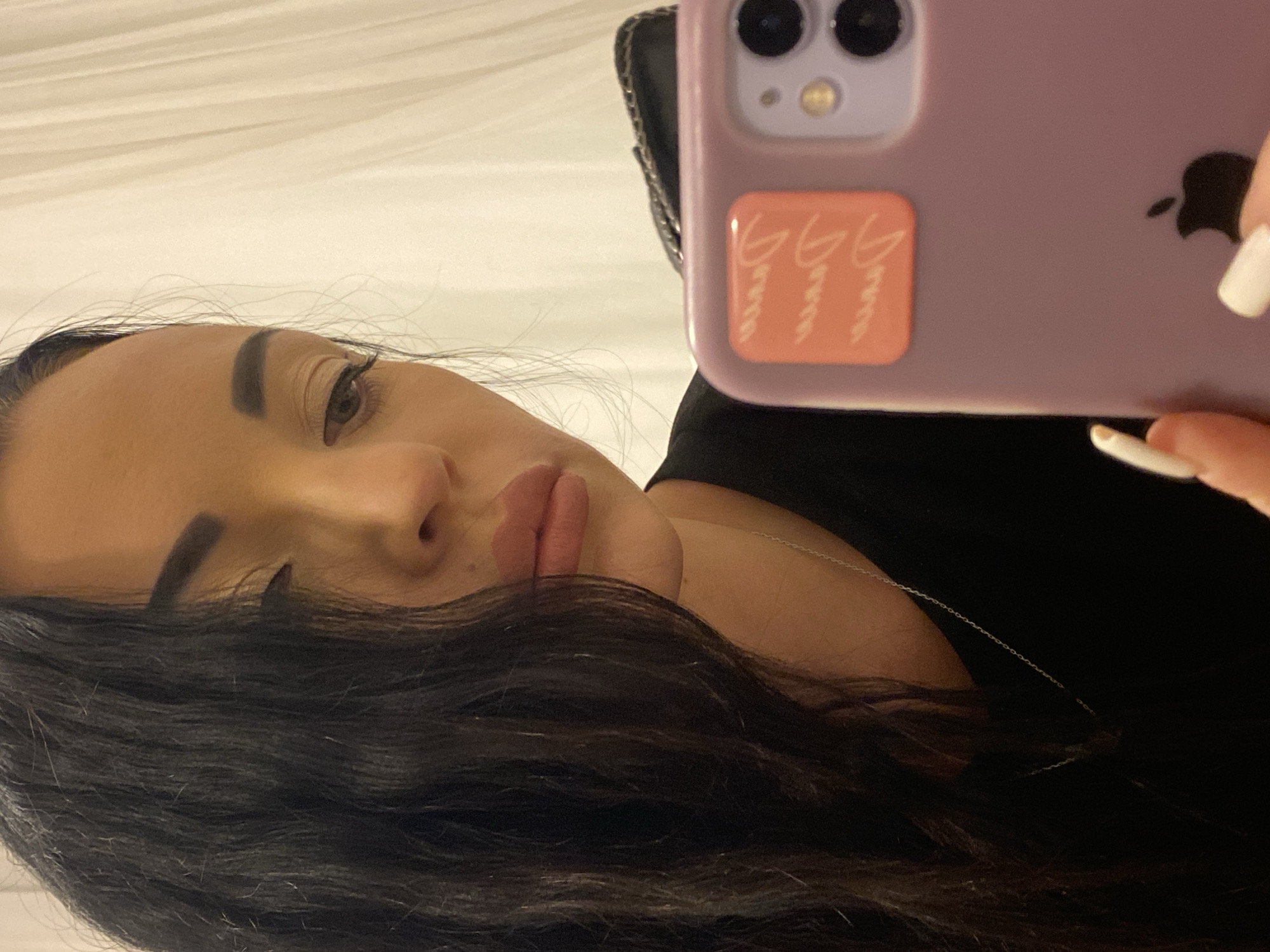 A medium shot, waist-up, selfie photograph shows a woman in Sharm El Sheikh, Egypt, holding up a purple iPhone to take her picture. She has long, dark, wavy hair that falls over her left shoulder and frames the left side of her face. Her right eye is visible, looking downwards, with dark mascara and eyeliner. Her eyebrows are dark and arched. Her lips are full and painted with a light pink lipstick. She is wearing a black t-shirt and a delicate silver necklace. The phone case has two small, square, orange stickers with white cursive writing that appears to say "Dior" repeated. The background is a blurred white curtain or drape. The lighting suggests an indoor setting, possibly a hotel room or a dimly lit space. The woman's fingernails are painted white.