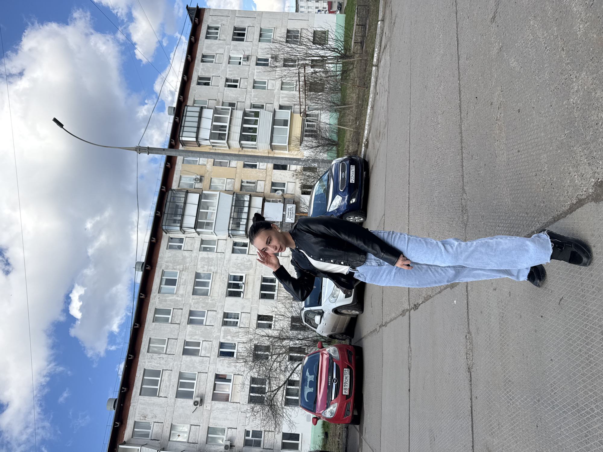 A young woman stands on an asphalt surface, seemingly a street or parking area, in Chaikovsky, Russia. She is wearing a black leather jacket, a white t-shirt, light blue jeans, and black shoes. Her hair is pulled back. She is looking at the camera and touching her forehead with her right hand. Behind her are three parked cars: a red Opel, a white car, and a blue Kia. A multi-story apartment building with a dilapidated facade and numerous windows is visible in the background. A street lamp is adjacent to the building. The sky above is partly cloudy with patches of blue.