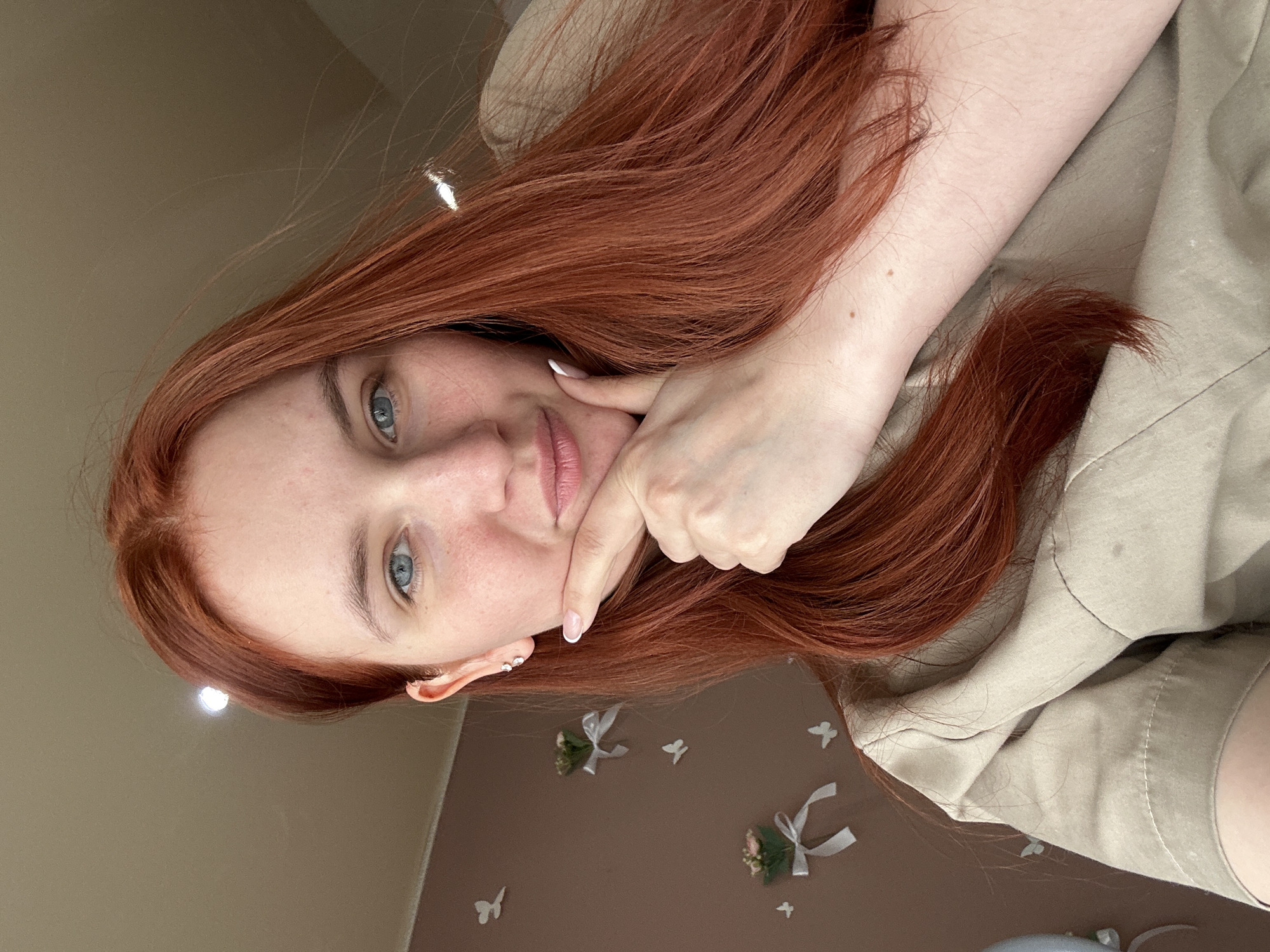 A young woman with long, vibrant red hair and striking blue eyes looks directly at the viewer in this indoor portrait, taken in Chaikovsky, Russia. Her fair skin shows a scattering of light freckles, and she has a gentle, subtle smile on her lips. She is seen resting her left hand (from the viewer's perspective) against her cheek and jawline, with her fingertips, adorned with white nail polish, lightly touching her skin.
She is wearing a light-colored, possibly beige or light green, long-sleeved top or shirt. Her auburn hair cascades around her shoulders and across her chest. The background features a two-toned wall; the upper section is a plain light tan or beige, while the lower section is a darker brown. This darker wall is decorated with several small, white butterfly cutouts and miniature floral arrangements tied with white ribbons, reminiscent of wedding favors or celebratory decorations. The lighting appears soft and ambient, with a bright reflection on the upper wall and in her hair, suggesting an overhead light source. No visible text is present in the image.