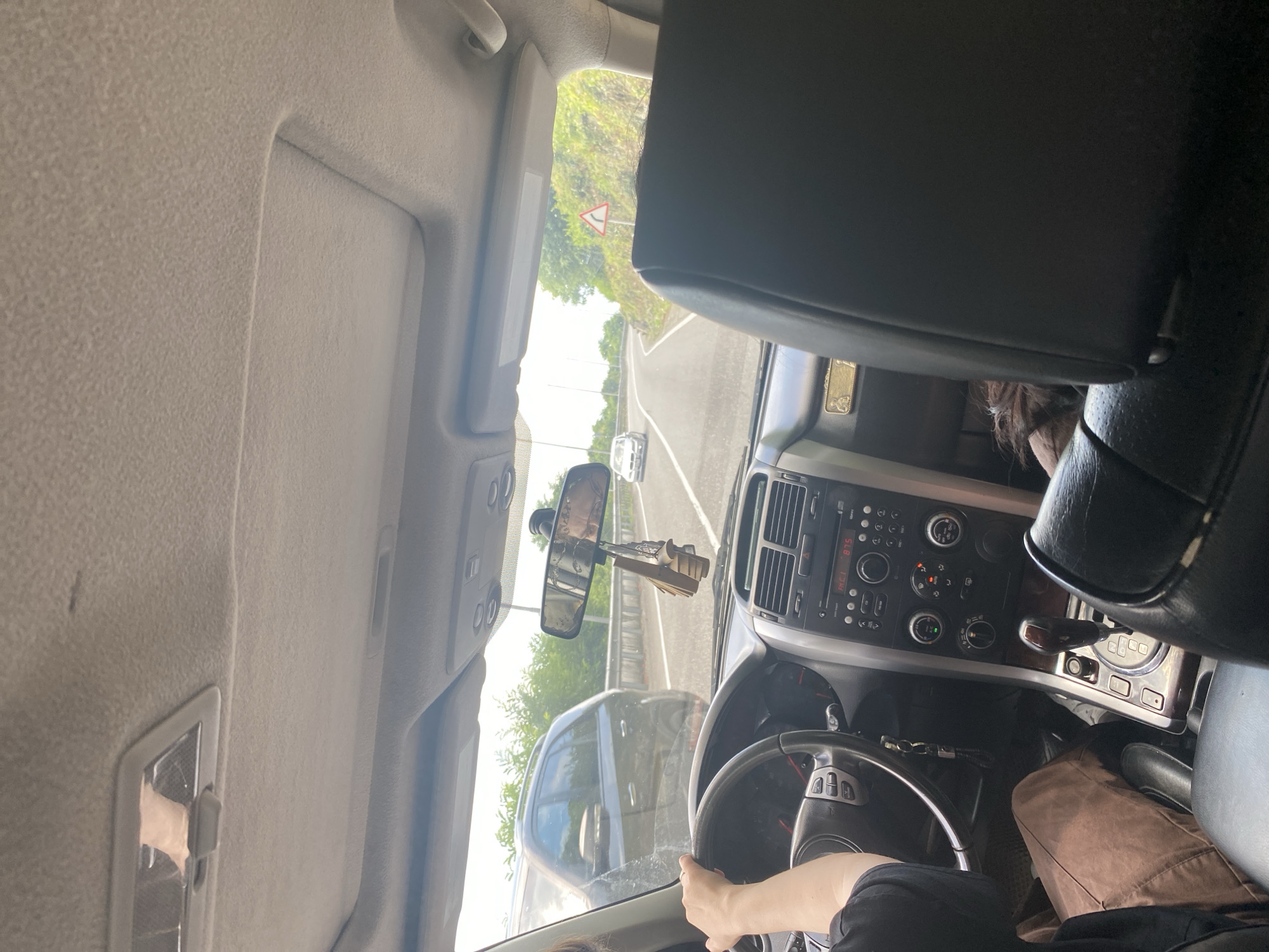 A first-person perspective shot from inside a vehicle, looking forward through the windshield. The driver's hands are on the steering wheel, and the passenger's head is partially visible in the background. The car's dashboard is in focus, displaying a radio and gear shift. Outside, a road winds through a green, hilly landscape. Another car is visible on the road ahead, and a third car is driving in the opposite lane. The sky is bright and overcast. The scene is set on a road in an unknown location.