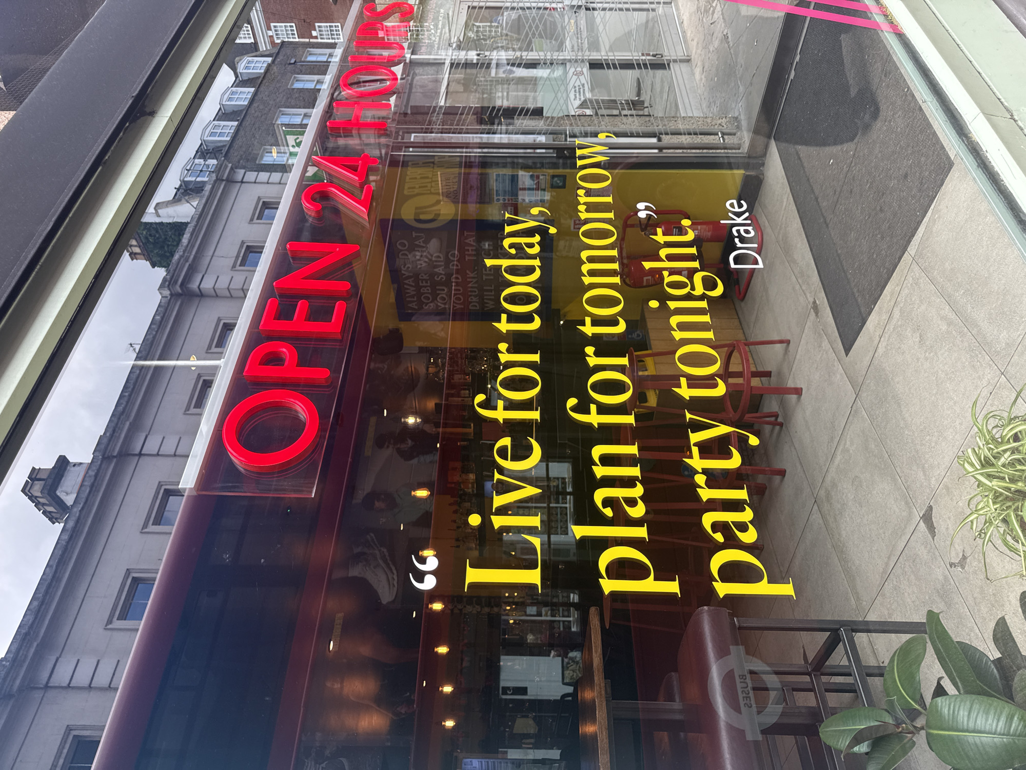 The image displays the glass facade of a commercial establishment, likely a bar or restaurant, in an urban setting, consistent with London, United Kingdom. Prominently featured on the window are large, glowing red neon letters spelling "OPEN 24 HOURS". Below this, a significant portion of the glass is adorned with large yellow lettering presenting a quote: "Live for today, plan for tomorrow, party tonight." This quote is enclosed in white quotation marks, with "Drake" attributed below it in white text.
Reflected in the glass are details of the surrounding environment, including a multi-story building with grey stonework, numerous windows (some bay-style), and a partial view of an overcast sky. A paved sidewalk and a dark grey doormat are also visible in the reflection at the bottom.
Through the transparent sections of the window, the interior of the venue is visible, showing red bar stools, a brown leather bench, and faint outlines of what appear to be shelves stocked with bottles. A red fire extinguisher is also discernible inside. Potted plants, one on the exterior pavement and another within the establishment, add some greenery.
Additional, smaller white text is visible on the glass, partially obscured by internal reflections, which reads: "ALWAYS DO SOBER WHAT YOU SAID YOU'D DO DRUNK. THAT WILL TEACH". Faintly visible from the OCR is "POLITE NOTICS", likely part of a small notice. The overall scene suggests a vibrant establishment geared towards entertainment and social gatherings, operating around the clock, with a theme of seizing the moment. The time of day appears to be daytime, under cloudy skies.