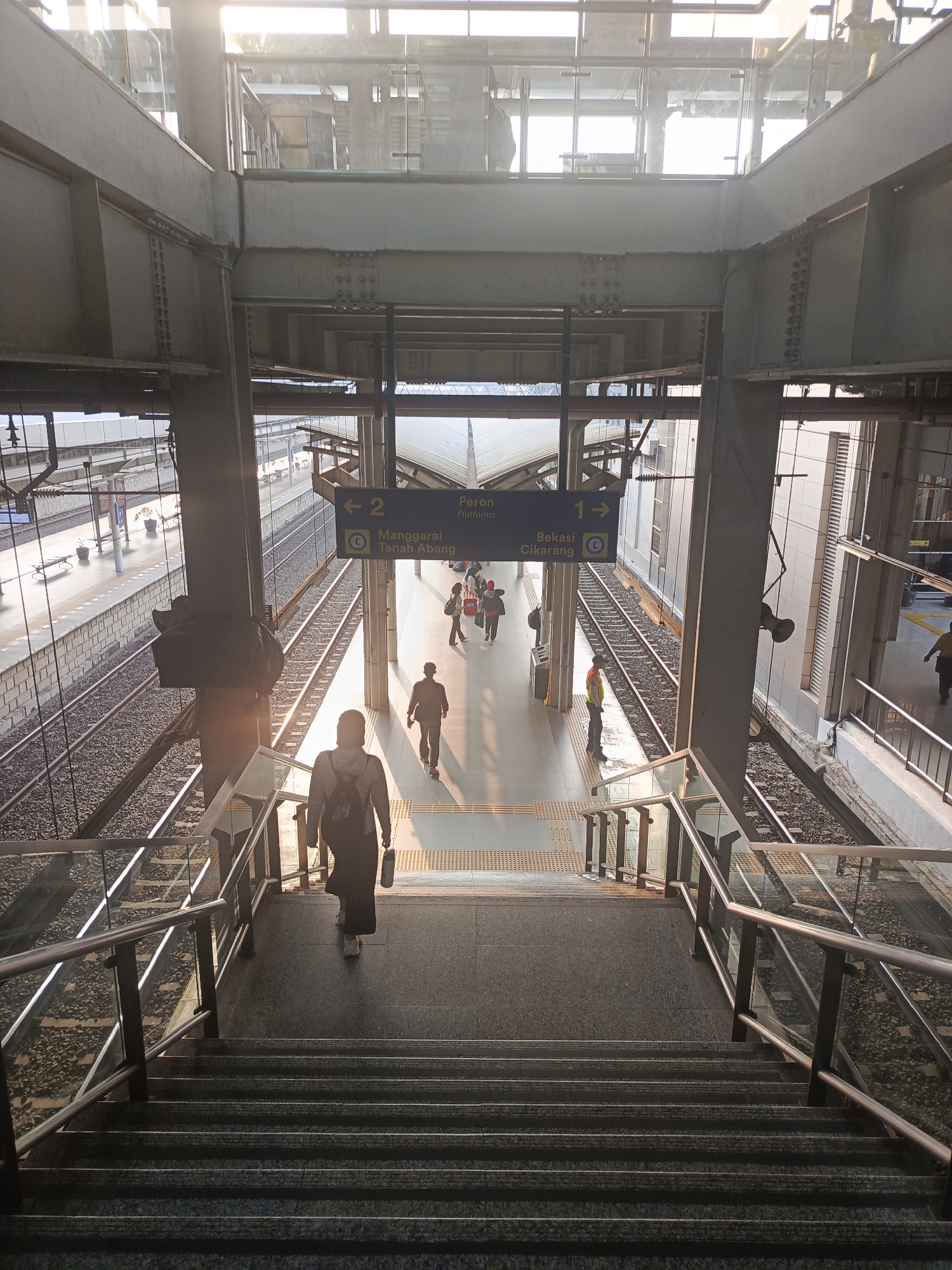 The image captures an interior view of a modern train station concourse in East Jakarta, Indonesia, during daylight hours, possibly late afternoon given the warm, directional lighting. The perspective is from a high vantage point, looking down a wide, long staircase with metallic railings and glass panels. The staircase leads to a concourse area with multiple railway tracks and platforms visible.
In the foreground, a lone individual wearing a dark hijab and a backpack is descending the stairs. Another individual, a man in a dark jacket, is walking away from the camera further down the concourse. Several other figures, appearing to be commuters, are visible on the platforms and in the distance, some walking and others congregating. A worker wearing a high-visibility orange vest is standing near the edge of a platform.
The environment is characterized by industrial architecture, featuring exposed steel beams, concrete structures, and elevated walkways. Overhead, glass panels allow natural light to flood the space, creating bright areas and casting long shadows. Signage in Indonesian, indicating train destinations like "Manggaraï Tanah Abang" and "Bekasi Cikarang," is visible above the platforms. The railway tracks are laid on a bed of ballast, and the platforms are equipped with tactile paving for the visually impaired. The overall scene suggests a busy, functional transportation hub.