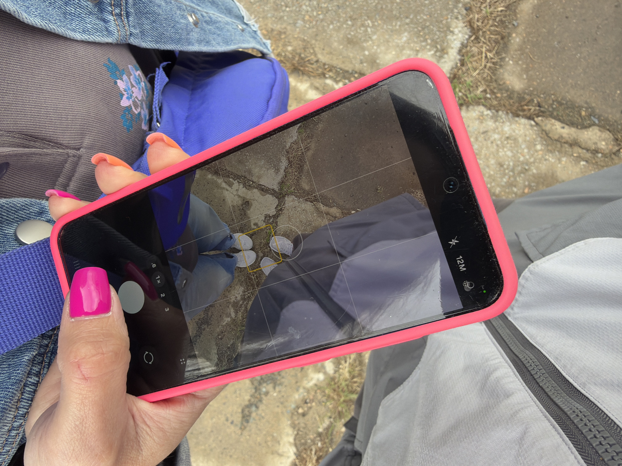 A first-person perspective image shows a person holding a smartphone in a bright neon pink protective case, actively using its camera function. The phone screen displays the camera interface with grid lines, zoom options (.6, 1x, 2, 3), a "12 M" indicator for resolution, and icons for flash/night mode. Reflected on the screen, a pair of white sneakers or shoes are visible, framed by yellow outlines, indicating they are the subject of the photograph.
The hand holding the phone has brightly painted fingernails, with the thumb featuring a solid hot pink polish and other visible fingers displaying a pink-to-orange gradient. The person is dressed in a blue denim jacket worn over a grey garment with blue and purple floral embroidery. A bright blue strap, possibly from a bag, is also visible. Part of a grey pair of pants or trousers can be seen in the lower right corner.
The background shows an outdoor setting with a rough concrete or paved ground surface interspersed with patches of dry grass and dirt. The bright, natural lighting suggests it is daytime. This scene is set in Nizhneleninskoe, Russia, and captures the act of taking a picture of one's footwear and the immediate ground.