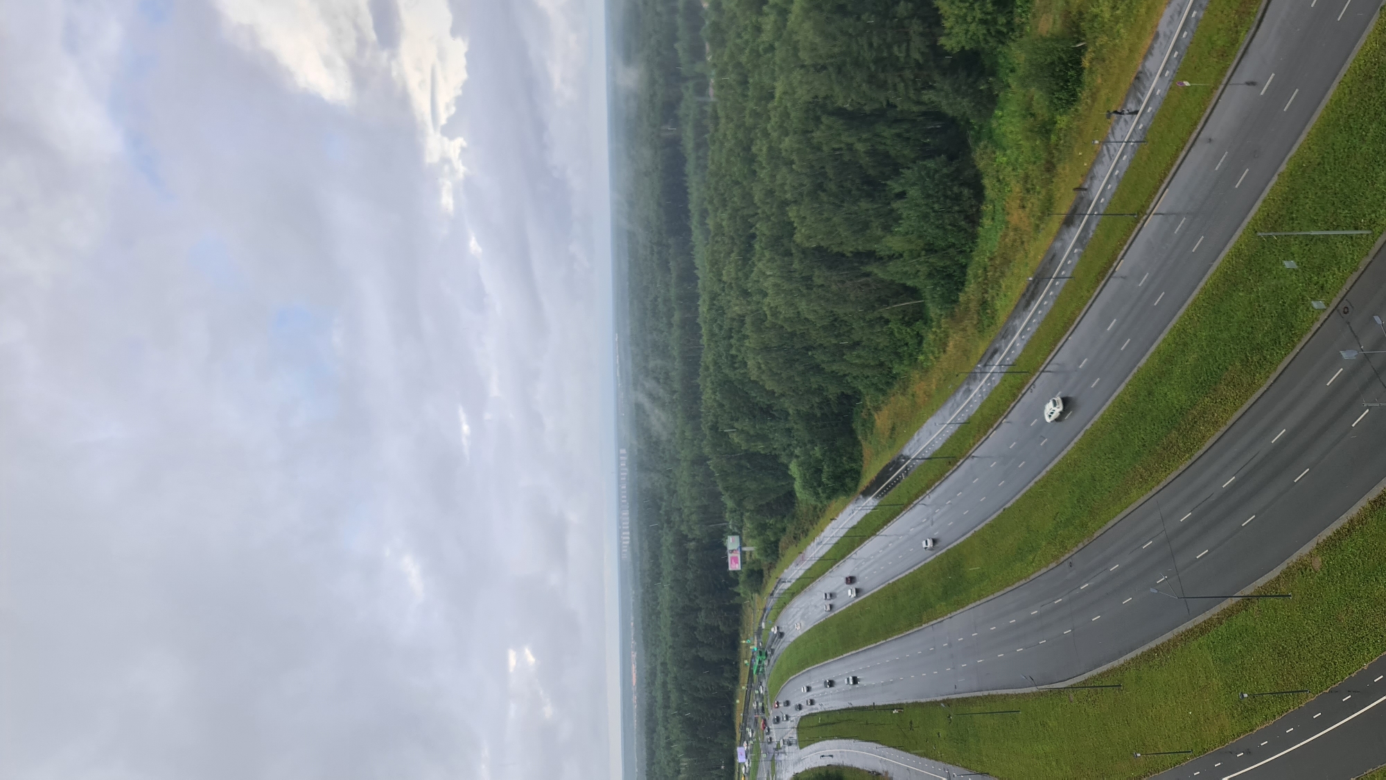 An aerial view captures a multi-lane highway on a cloudy day, with rain visible as streaks across the scene. The highway is bordered by green grassy verges and a dense forest of green trees. In the distance, a line of buildings or structures can be seen on the horizon. Several cars are visible on the highway, and a few pedestrians are present on a path alongside the road. The road surface is wet, indicating recent or ongoing rain. The location is the M5 Ural Highway near Chelyabinsk, Russia.