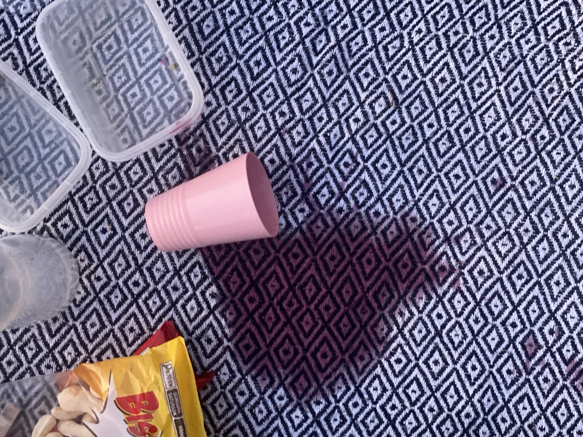 The image presents an overhead view of several items on a black and white geometric patterned fabric, likely a picnic blanket or tablecloth. The fabric features a repeating diamond motif.
In the upper left, two clear, rectangular plastic food storage containers with lids are visible, reflecting the underlying pattern. To their left and slightly below, a portion of another clear plastic cup or bottle can be seen.
A ribbed pink plastic cup lies on its side towards the center of the frame. A significant dark red or purple liquid spill, originating from the pink cup, spreads across the patterned fabric, creating a large, irregular stain that darkens the material in the central and lower-right sections of the image.
In the bottom-left corner, a yellow snack bag is partially visible. Prominently displayed text on the bag includes "Big" in red letters. Smaller white text indicates Brazilian nutritional warnings: "ALTO EM", "GORDURA SATURADA", and "SÓDIO". The bag appears to contain light-colored snacks, possibly nuts.
The scene suggests a casual eating or snack event, potentially a picnic or an informal gathering, interrupted by a spilled beverage. The lighting indicates it is daytime. No people are visible. The visible text on the snack bag confirms the context of São Paulo, Brazil.