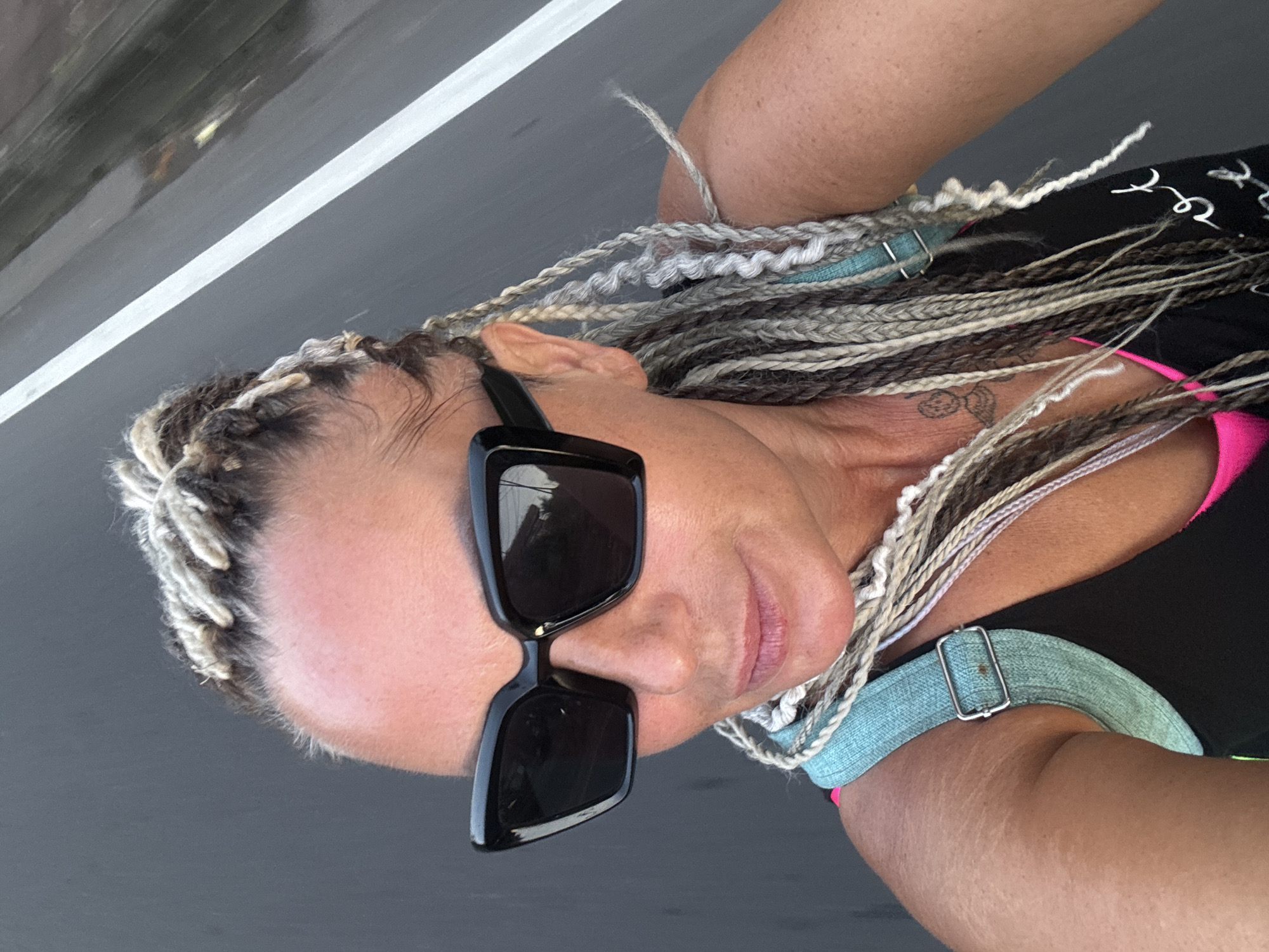 A woman with braided hair and large, black sunglasses is posing in a photograph taken outdoors. She is wearing a black tank top with a pink undershirt visible at the neckline. A light blue strap with a buckle is around her shoulder and arm. A small tattoo is visible on her upper chest. The background consists of a road with a white line marking the lanes. The exact location within Kuta Selatan is not discernible from the image.