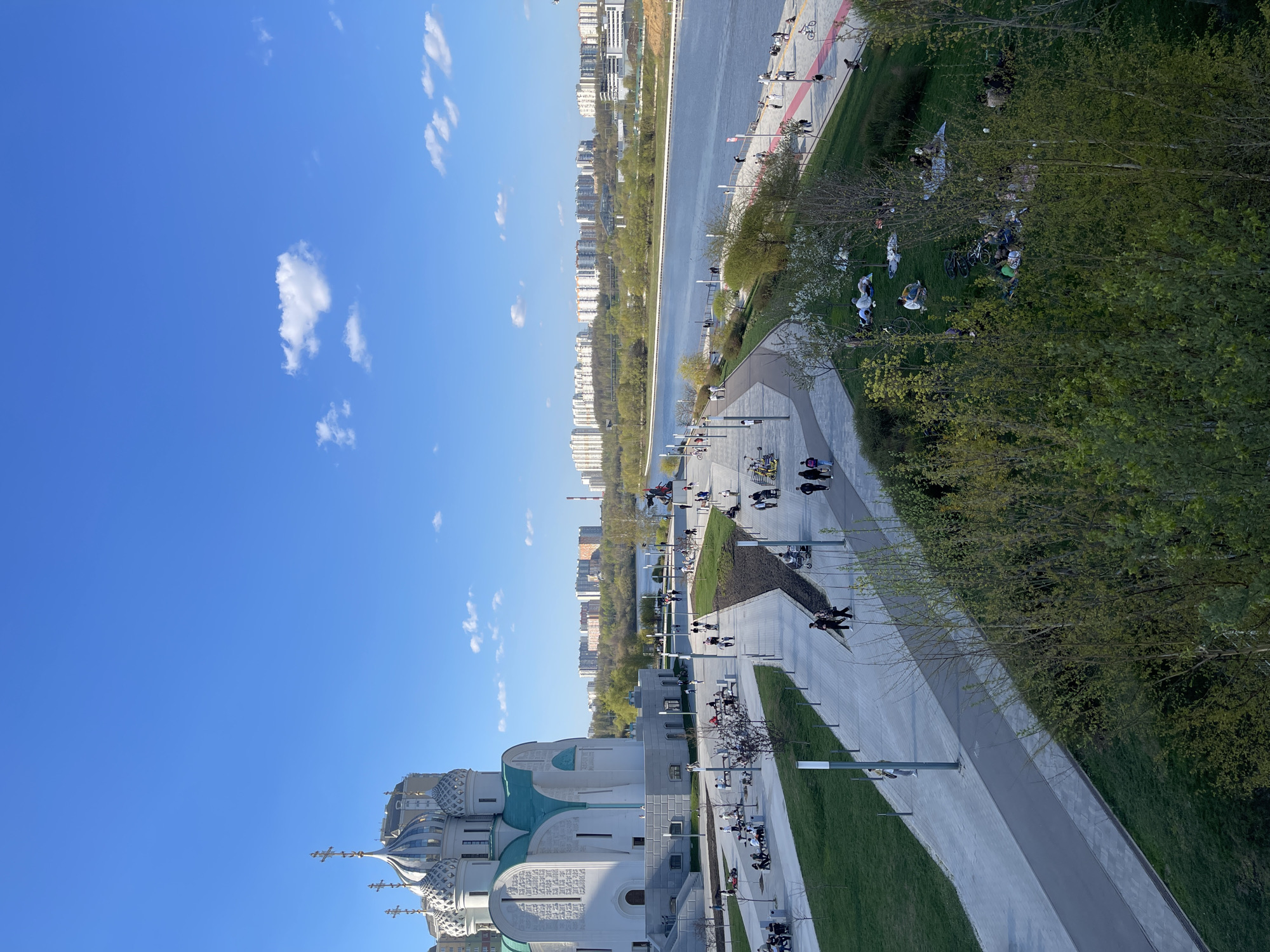 The image shows a view of Khimki, Russia. A white orthodox church with green and white domes occupies the lower left of the frame. A wide pedestrian walkway runs parallel to the river. People are walking, sitting, and biking along the walkway and in the adjacent green spaces. Trees line the riverbank, and apartment buildings are visible in the background across the river. The sky is blue with scattered clouds.